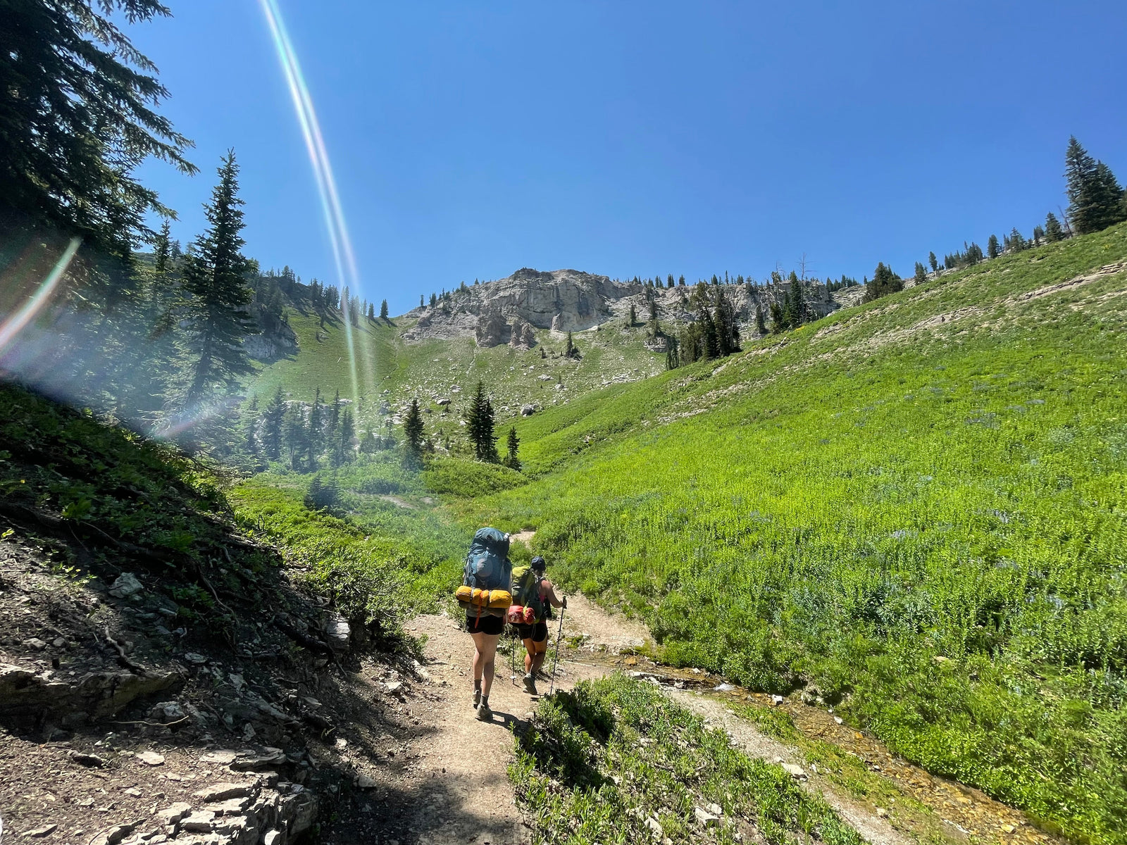 Two girls with backpacking gear hiking on a trail. There is bright blue sky and lots of green grass, it is a beautiful outdoor day.