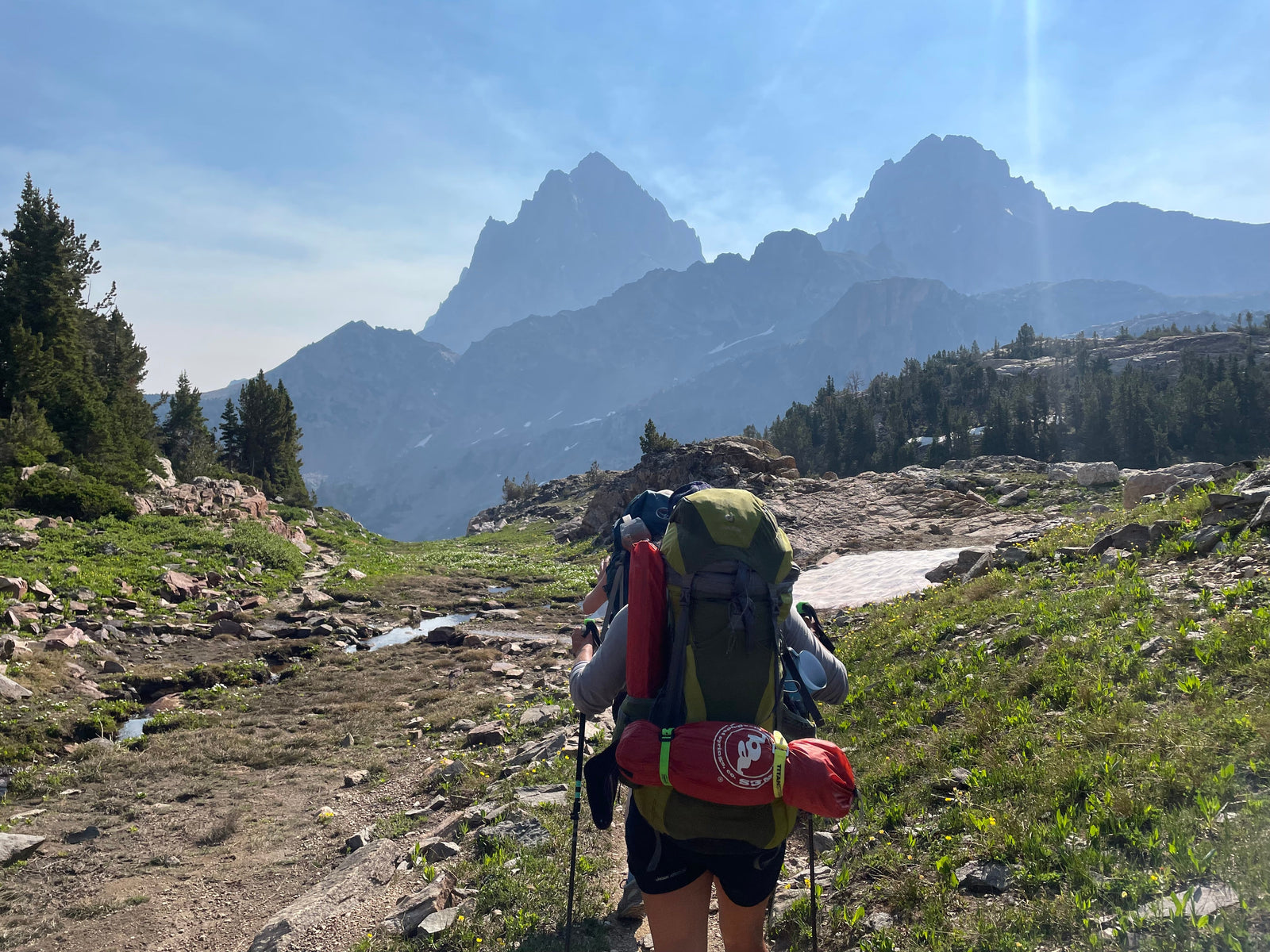 Looking at two girls hiking in the photo. They are walking towards a huge mountain range (the Tetons) in Wyoming on a beautiful sunny day. They are wearing the backpacking gear and appear to be on the trail for several days.