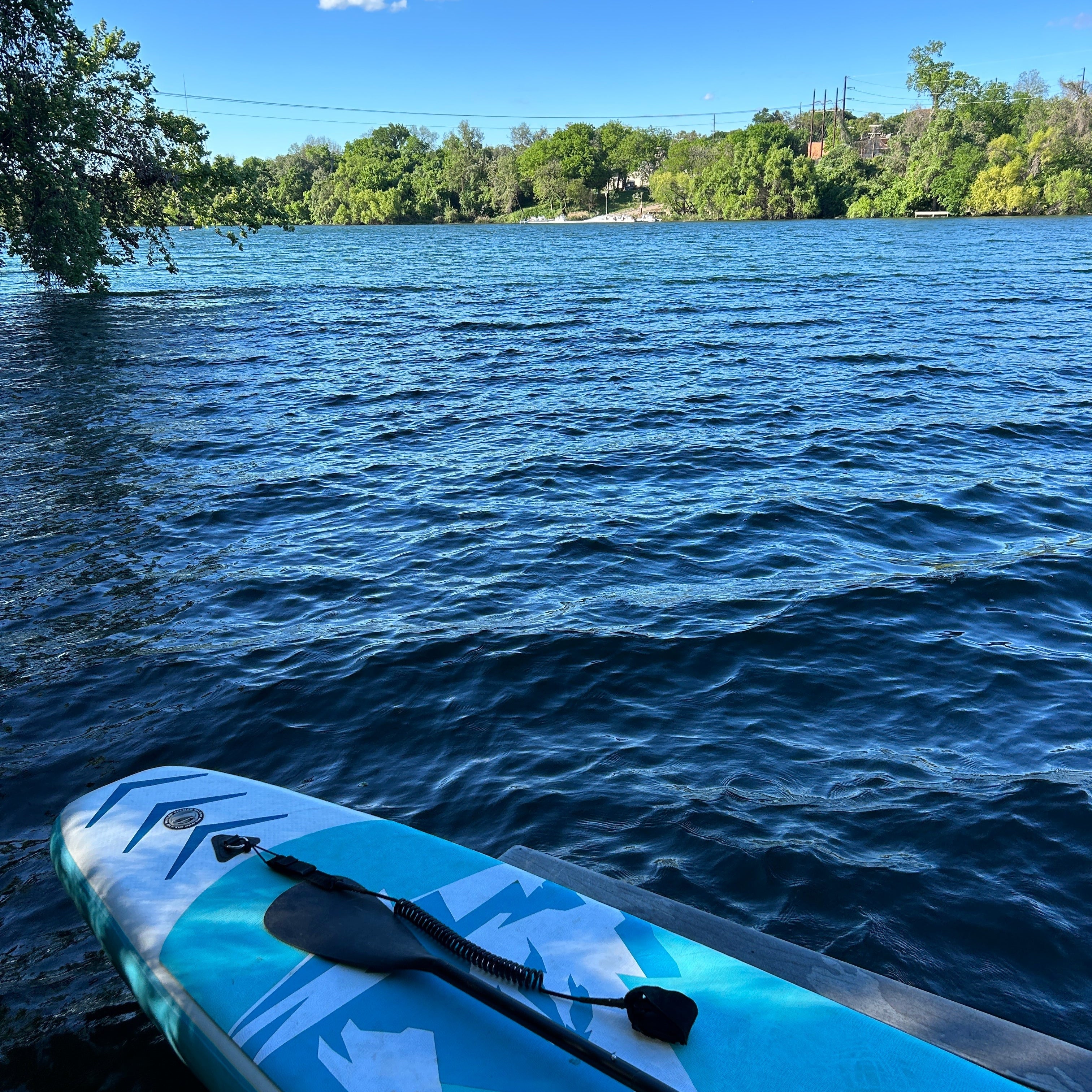 A blue paddleboard is laying on a wooden dock next to the water. It looks ready to go in the blue water. There is blue sky in the distance and the lake is surrounded by full green trees.