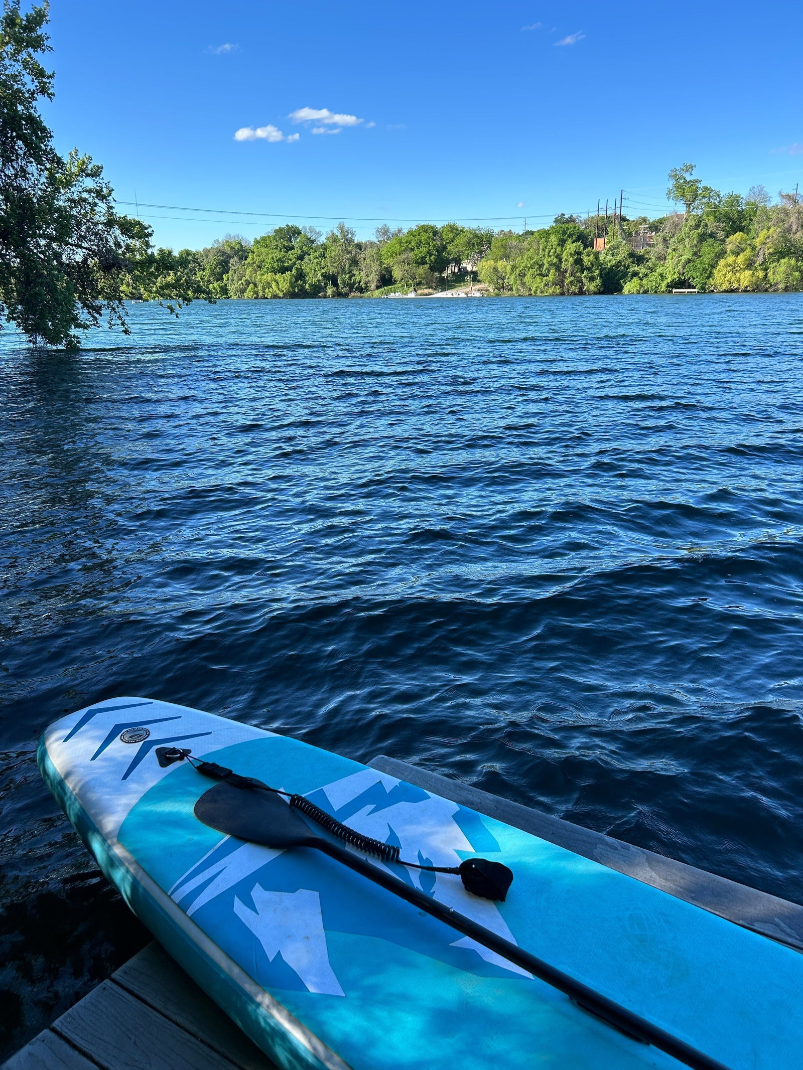 A blue paddleboard is laying on a wooden dock next to the water. It looks ready to go in the blue water. There is blue sky in the distance and the lake is surrounded by full green trees.