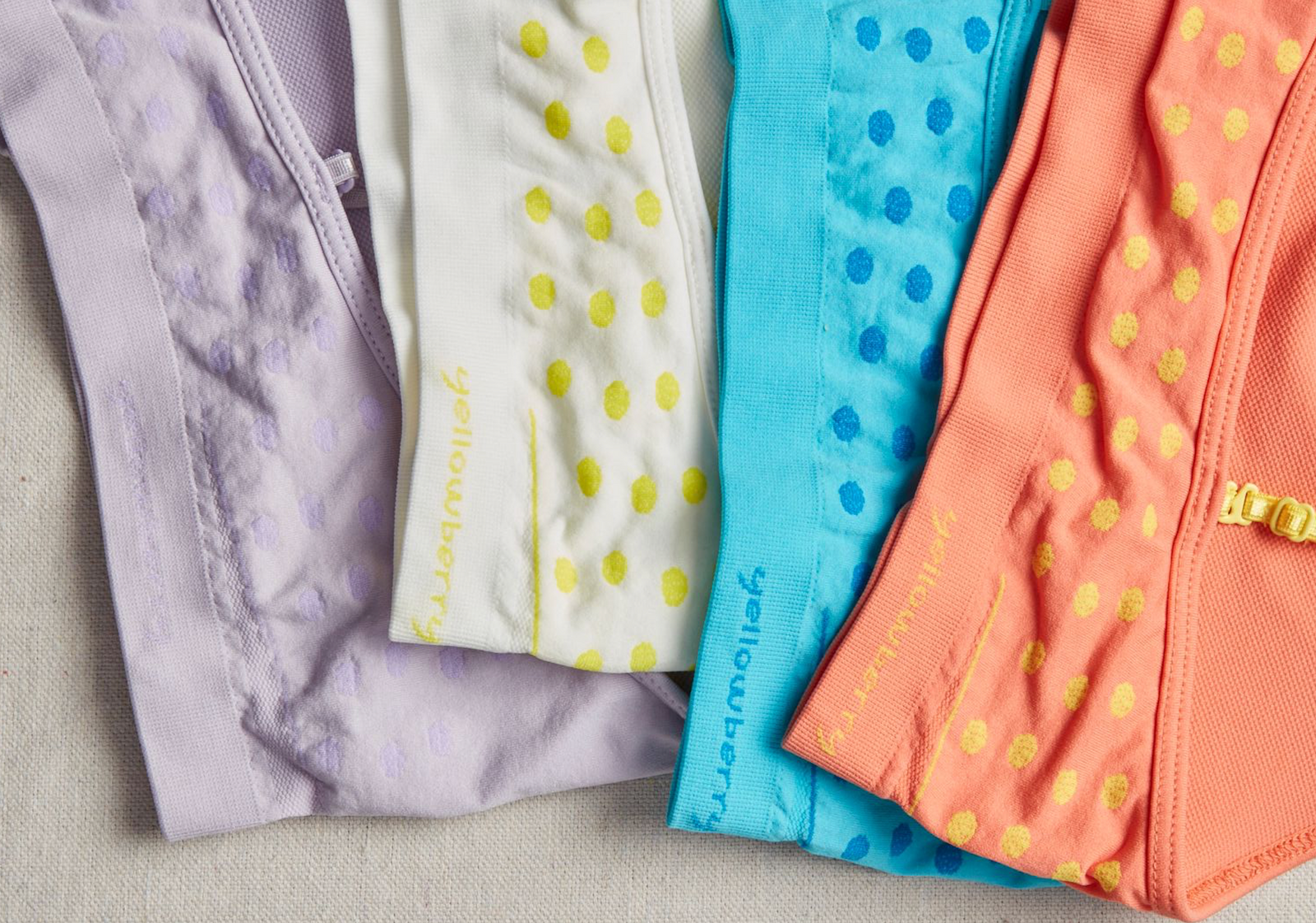 Four different colored seamless bras laying on a light colored linen background. One is lavendar, on is white, one is blue, and one is a coral color. Each has the Yellowberry name sewn into it.