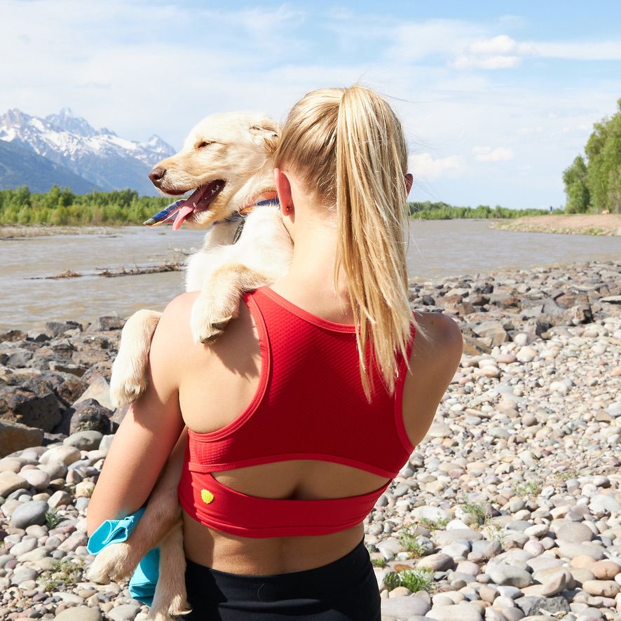 Girl holding an adorable labrador puppy, facing away from the camera along a stream. Her blone hair is in a ponytail, and she is wearing a red Yellowberry crop top