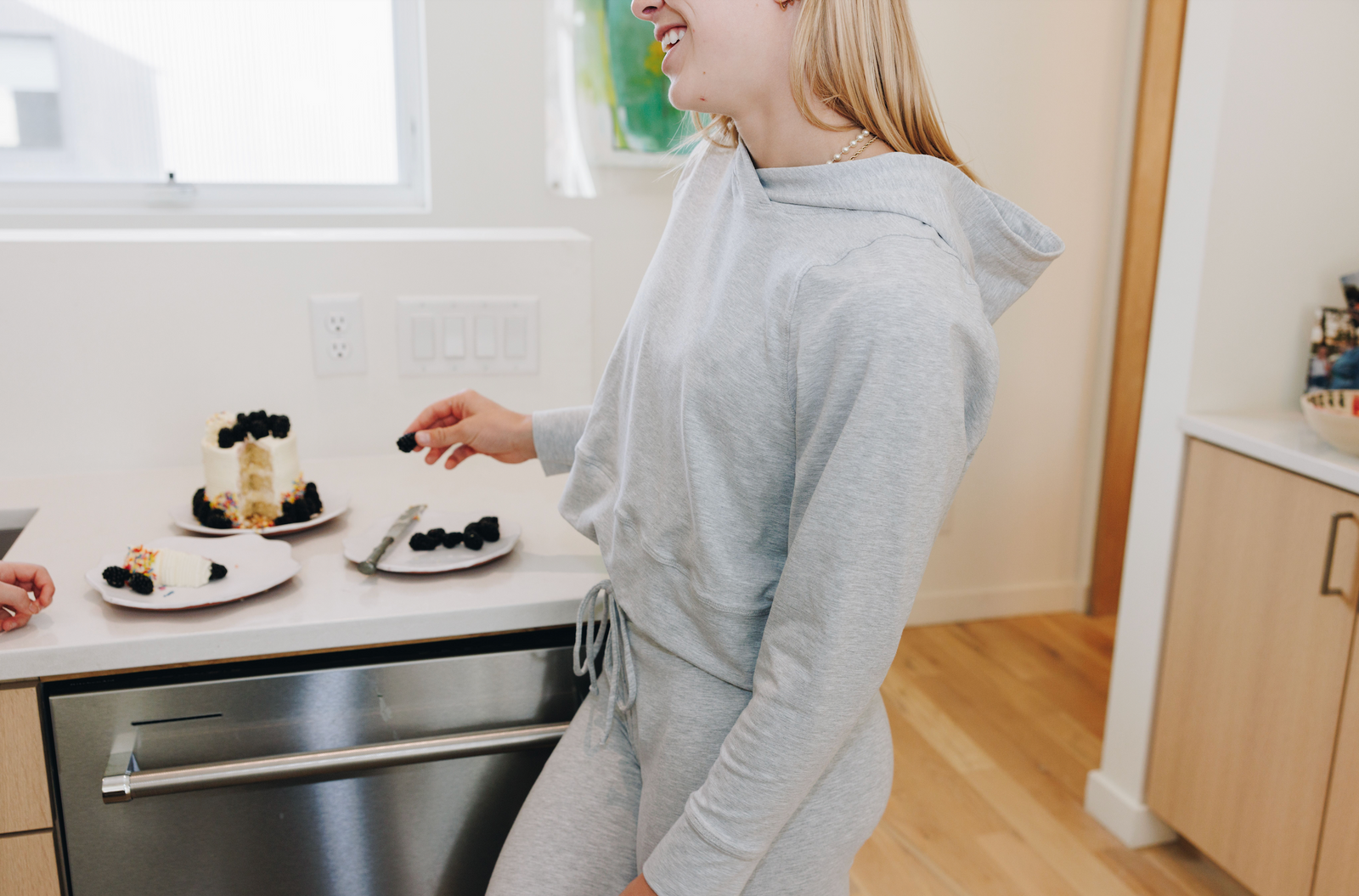 Girl smiling and wearing a grey hoodie. She is leaning against a counter and holding a piece of cake in her hand.