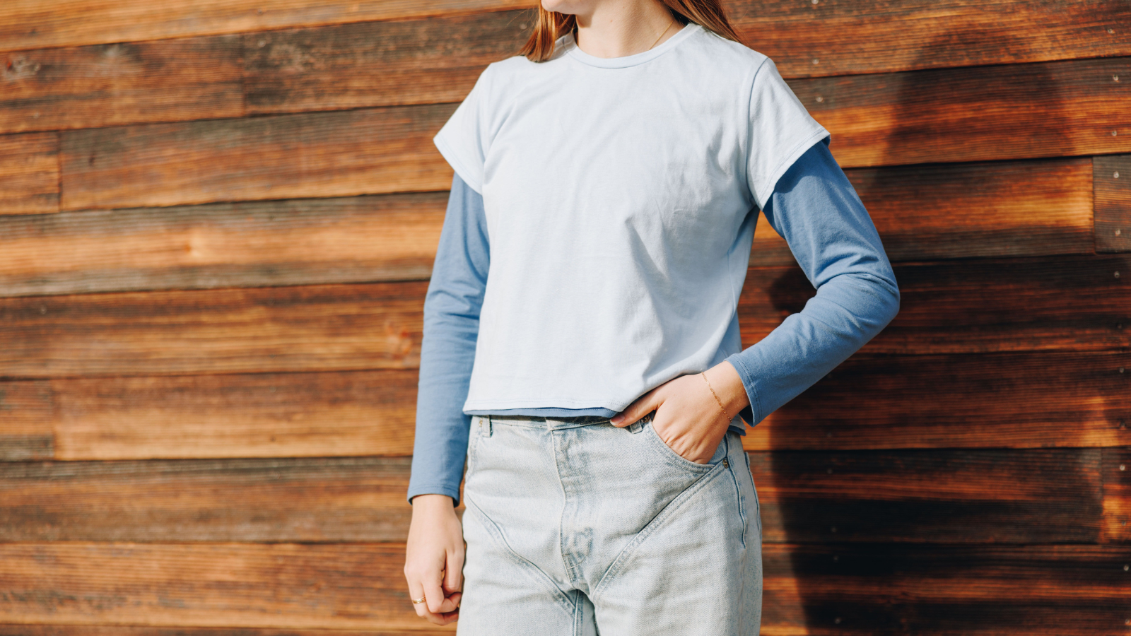 Girl wearing two layered blue t-shirts standing against a wodden wall with her hand in her pocket and face out of frame