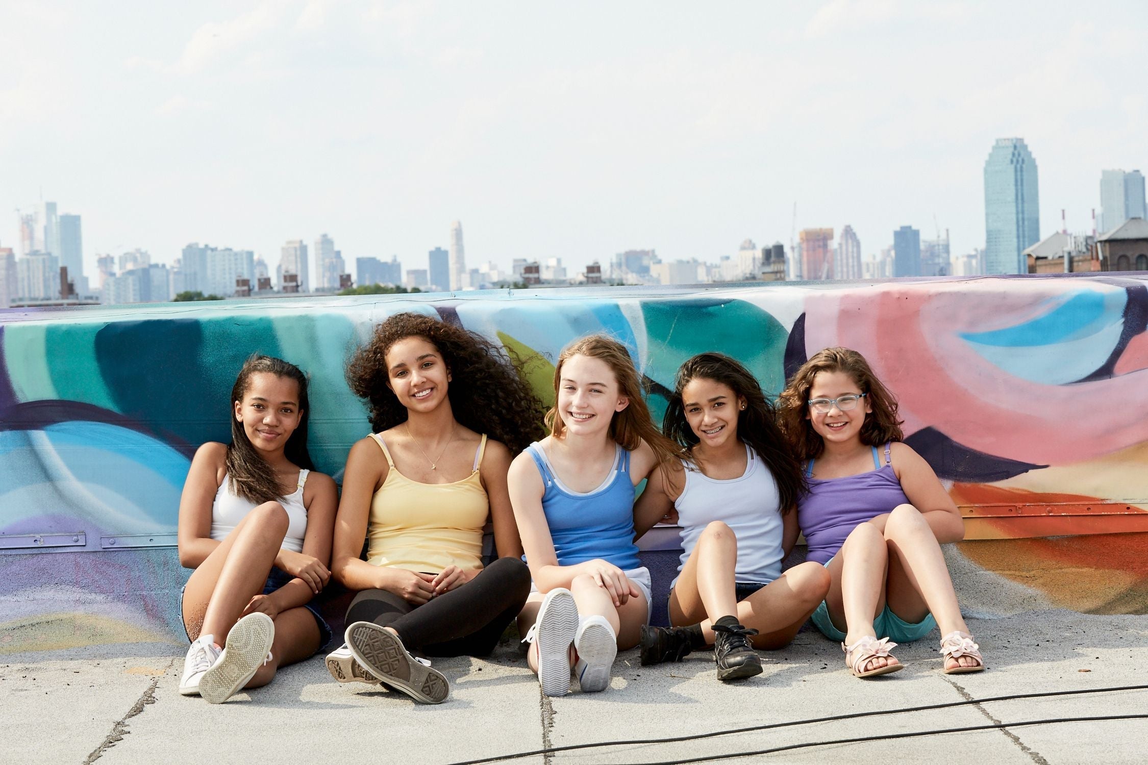 Five girls sitting on the ground, leaning against a colorfully painted wall. The city building, city-scape is behind them. They are all wearing different colored Yellowberry Camisoles, Tank Tops, and Lounge pieces.