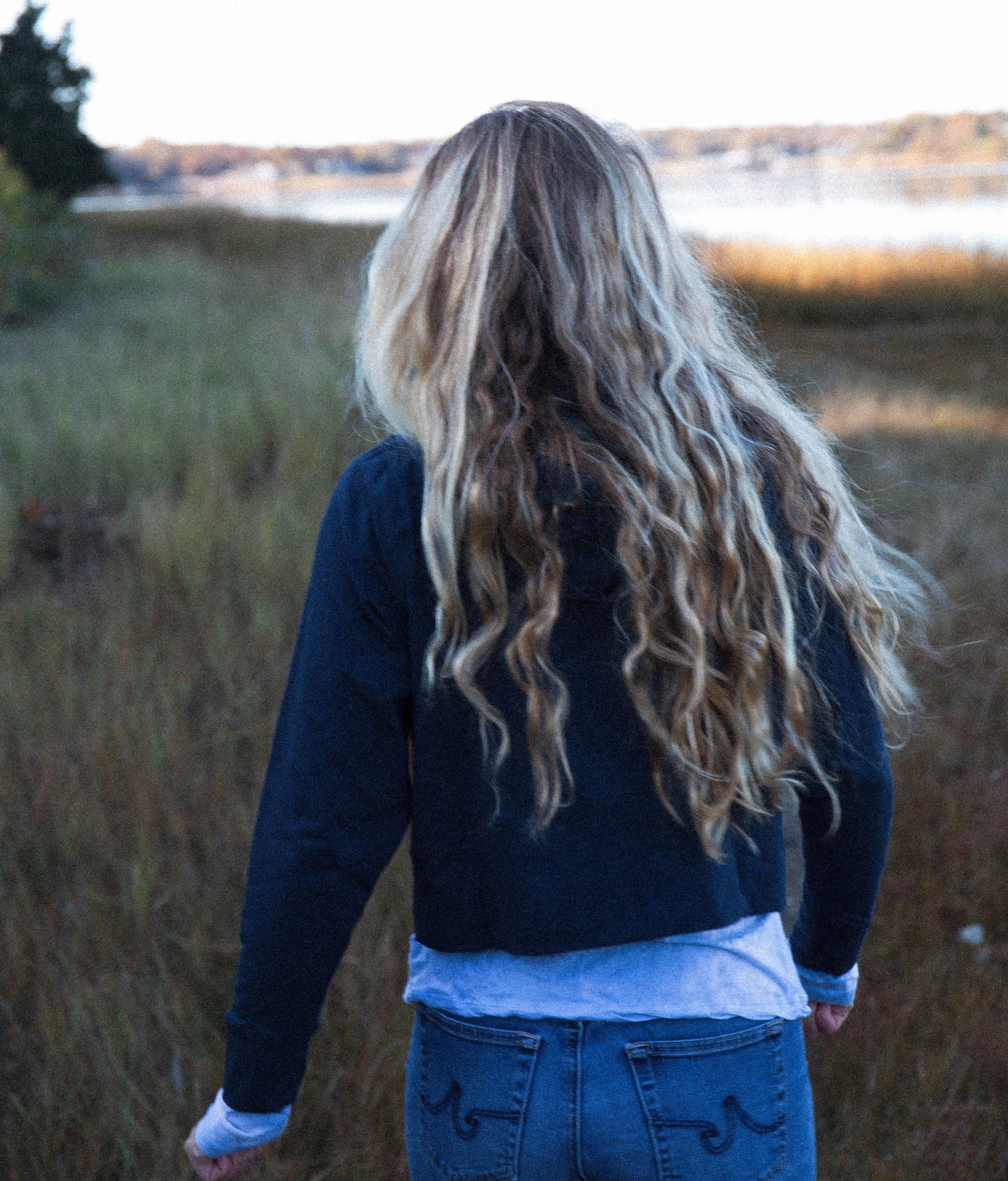 Girl with long blonde hair down her back, walking away from the camera in a meadow