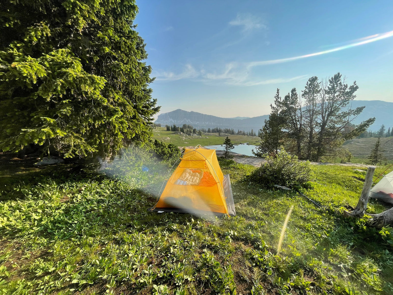 A Yellow tent is set up on the side of a hill overlooking a blue lake in the distance. It is surrounded by green grash and lush green mountain trees.