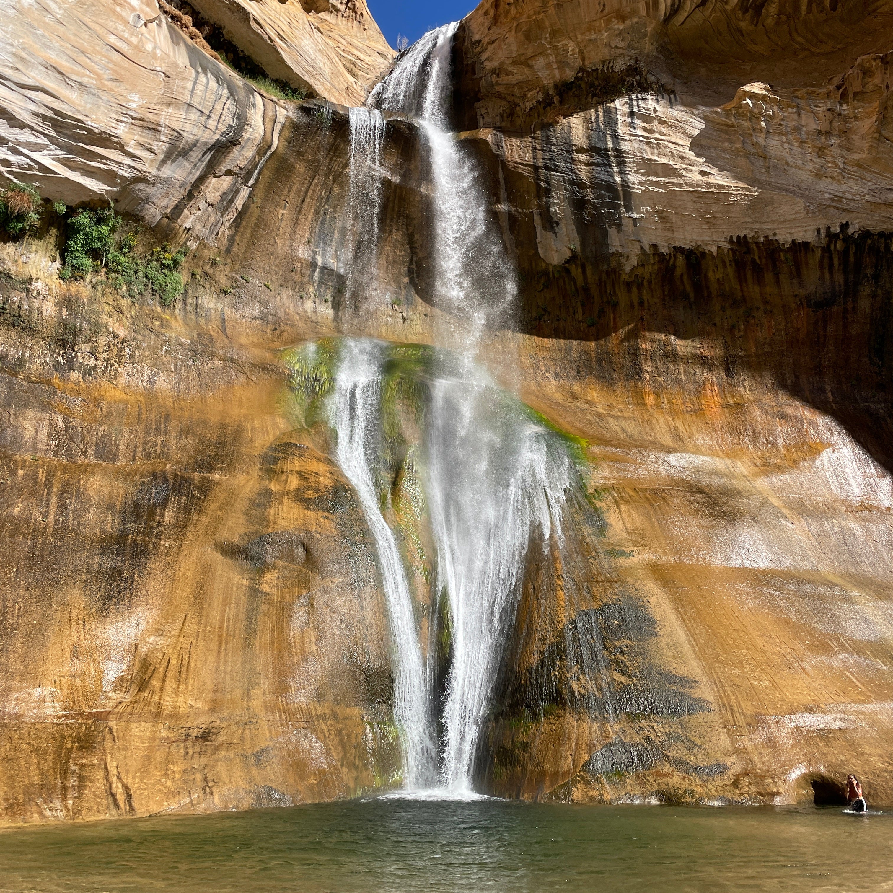 A gorgeous natural, red rock waterfall. It's massive, and comes into a small pool at the base. Background is more red rock and a small view of the blue sky.