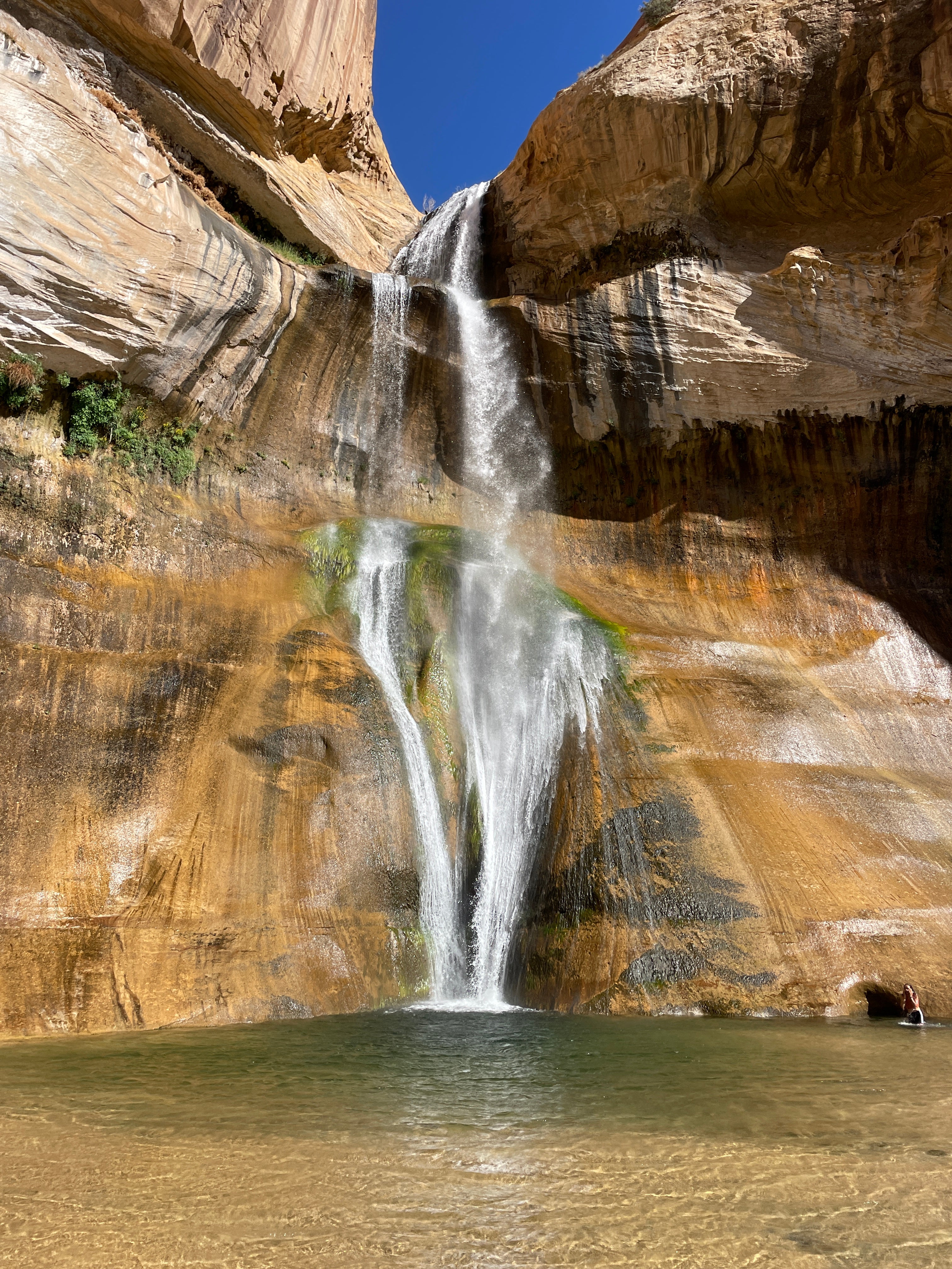 A gorgeous natural, red rock waterfall. It's massive, and comes into a small pool at the base. Background is more red rock and a small view of the blue sky.