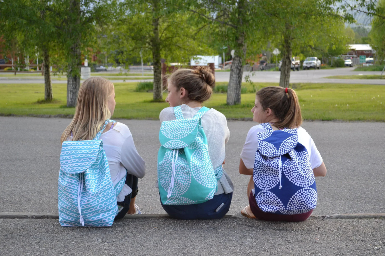 Three girls sitting on a curb. They are wearing t-shirts and jeans and all wearing colorful backpacks.