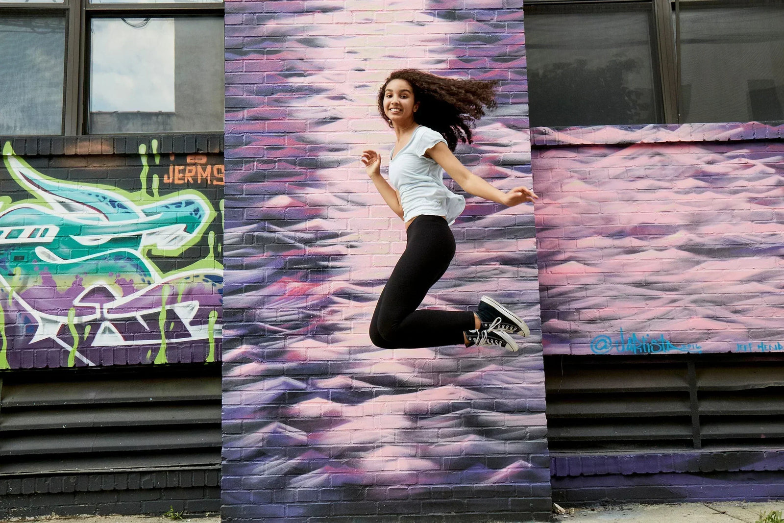 Girl jumping high above the ground. She looks like she is floating next to a colorful, painted mural on the building behind her. She is smiling!