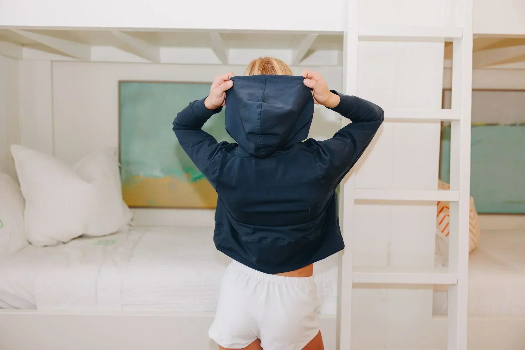 Girl pulling up her hood on the Yellowberry navy Downtown Hoodie she is wearing. She is facing away from the camera, and also wearing a pair of white Yellowberry Midtown Shorts. She is standing in front of a white bunk bed.