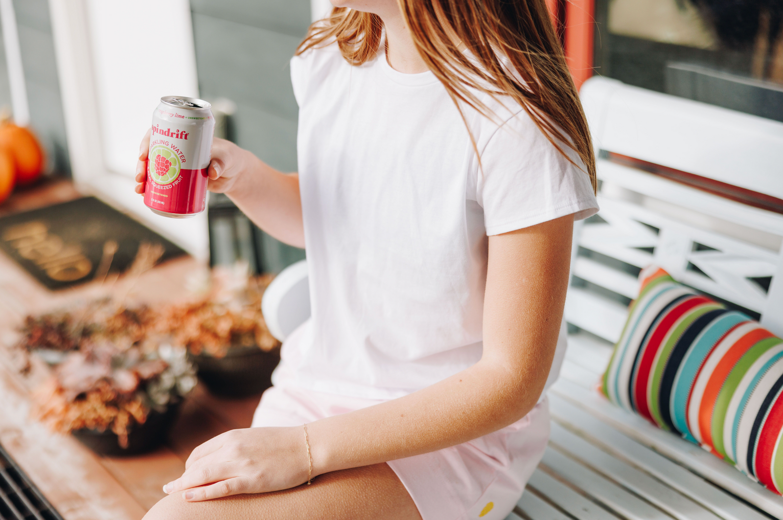 Girl sitting down on a bench in a white cotton t-shirt and pink shorts holding a drink in her hand.