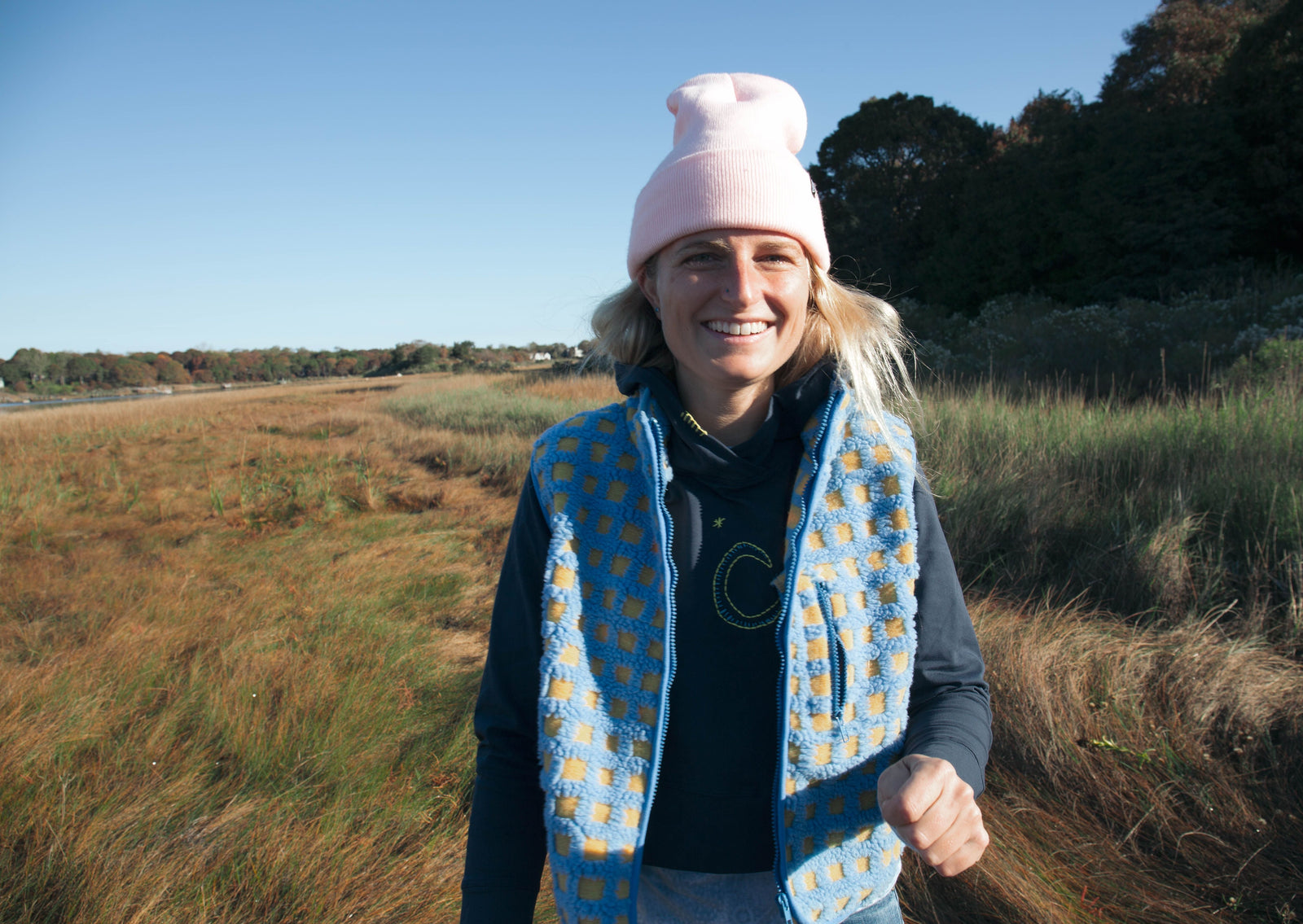 Girl smiling at the camera and standing in a field. She is wearing a pink hat with a navy, embroidered Yellowberry hoodie underneath a yellow and blue patterened vest. It is a beautiful sunny sky behind her.