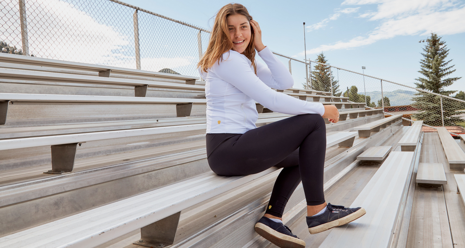 Girl is sitting on metal bleachers while wearing a white Yellowberry hoodie and black Yellowberry leggings. She is tucking her hair behind her ears with the blue sky behind her