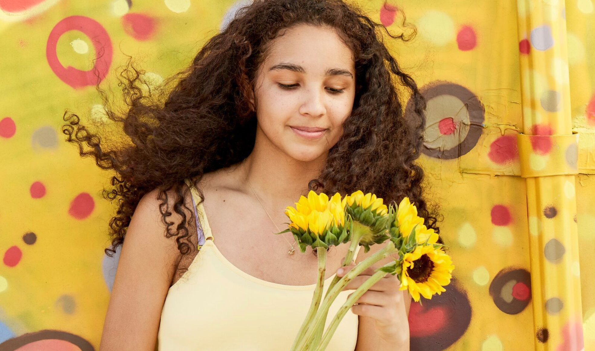 Girl is smiling down at the sunflowers in her hand, wearing a yellow cotton top, standing in front of a yellow themed mural