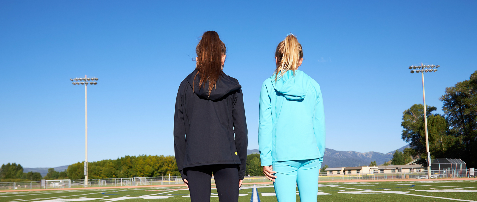 Two girls standing with their backs to the camera. One is in a black hoodie and the other is in an aqua blue hoodie. They face a blue morning sky
