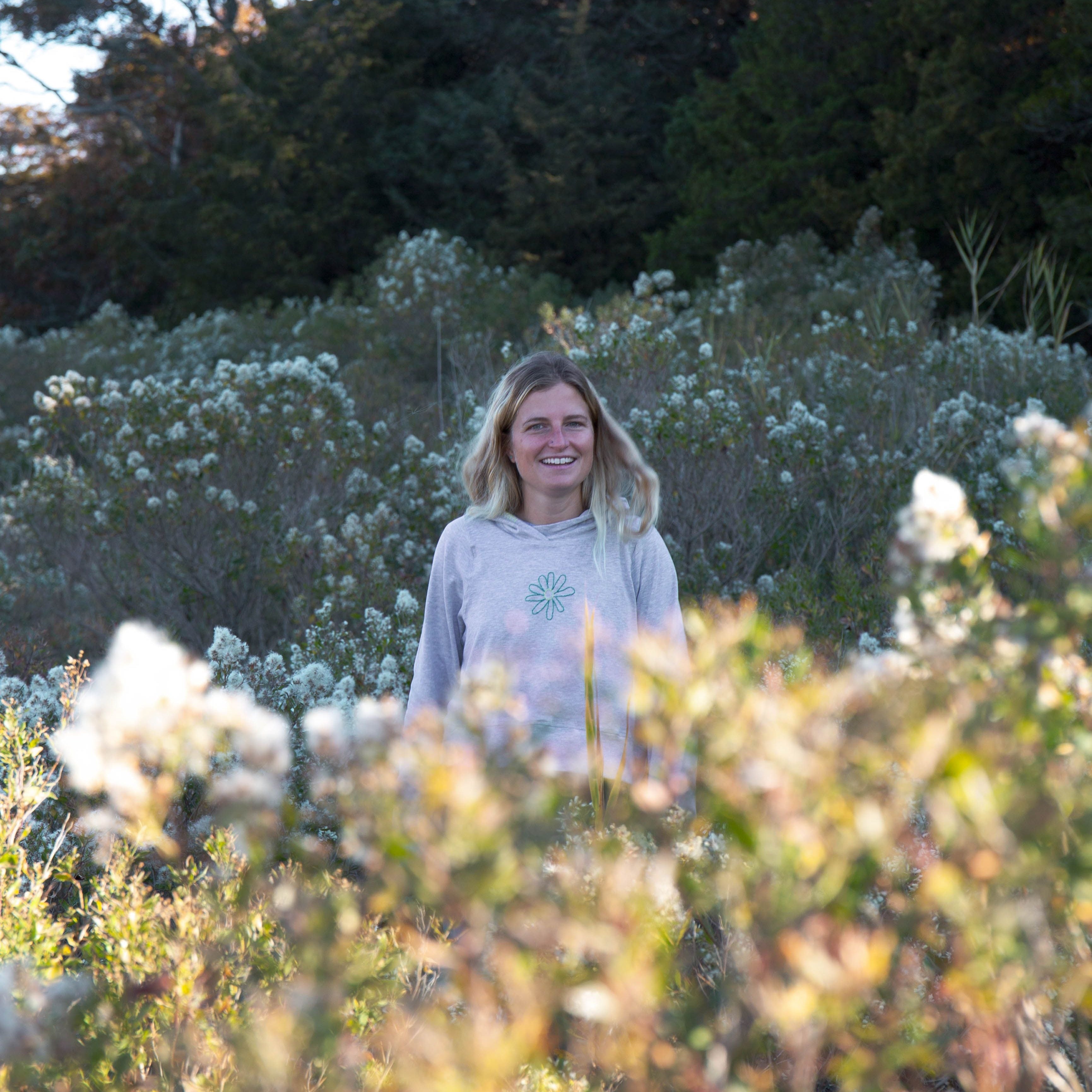 Girl standing behind flowers in a field. She is wearing a grey Yellowberry Downtown Hoodie and smiling at the camera.