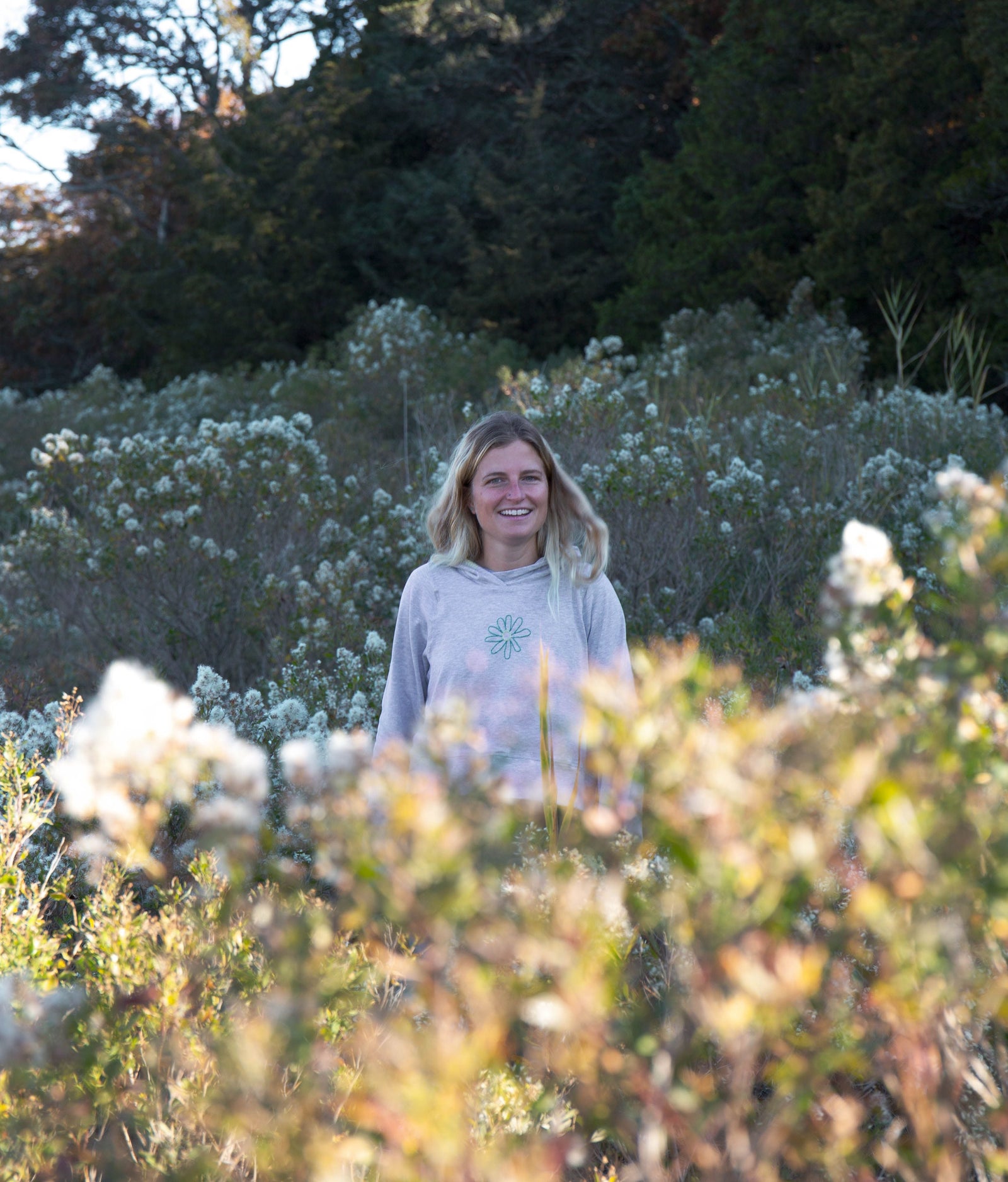 Girl standing behind flowers in a field. She is wearing a grey Yellowberry Downtown Hoodie and smiling at the camera.
