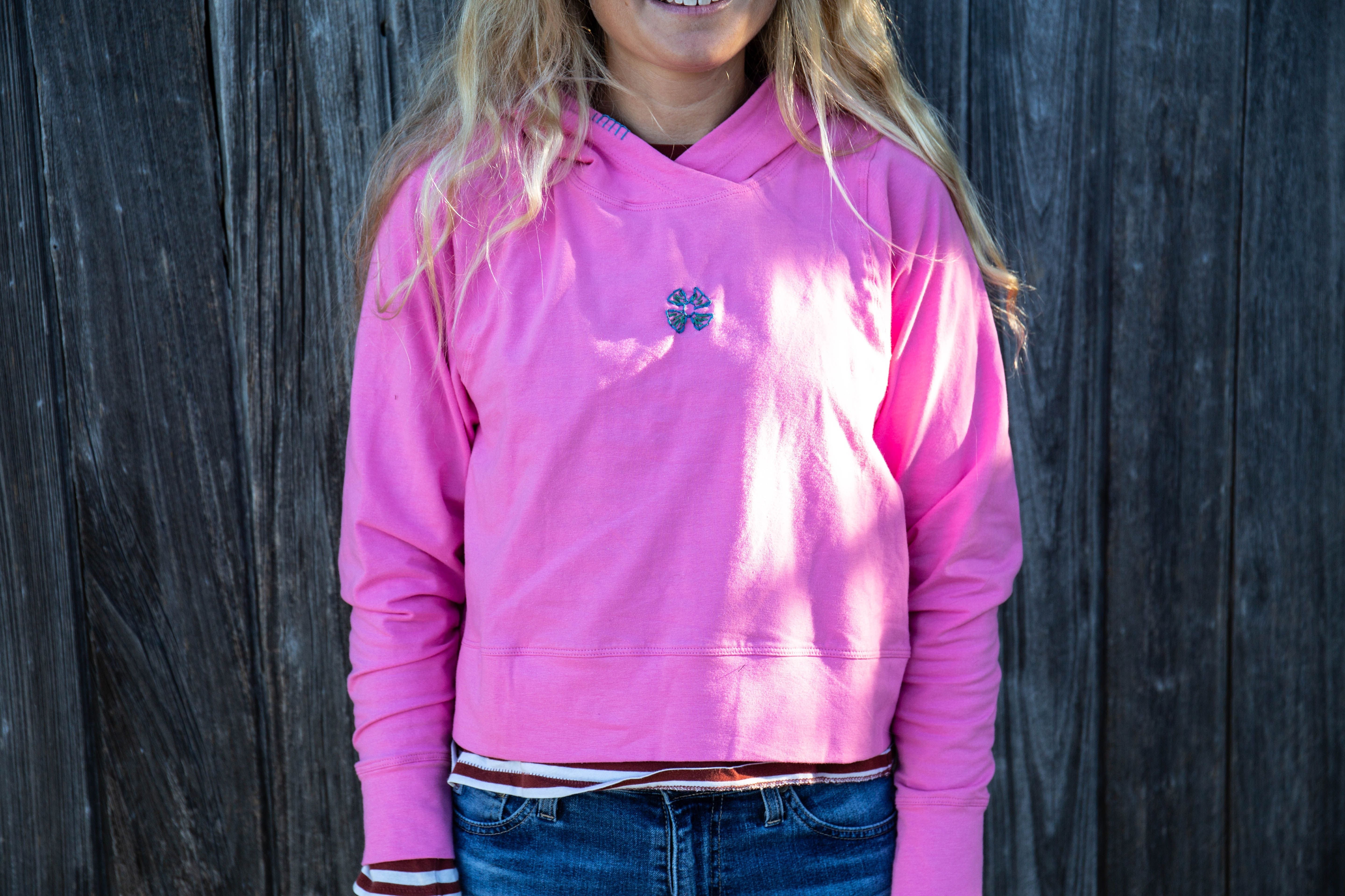 Girl wearing a bright pink hoodie with an embroidered deisgn on the front. She is standing in front of a wooden wall.