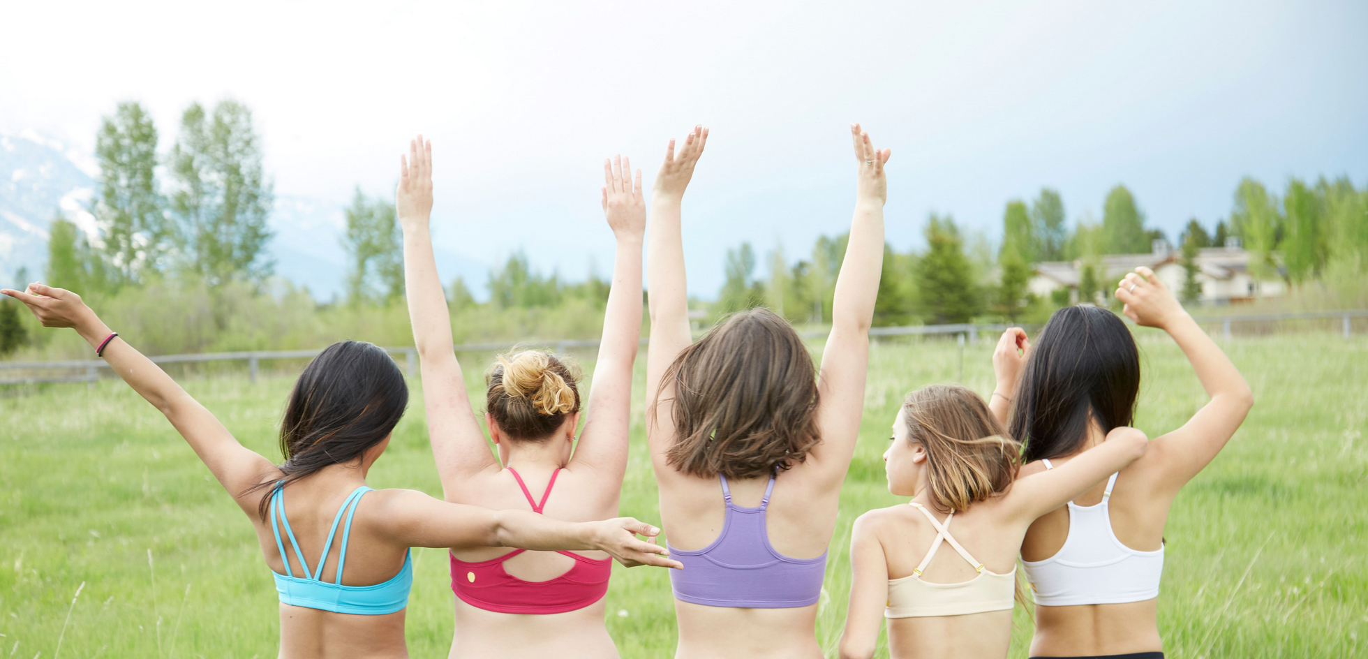Camera is behind 5 girls in colorful, youthful Yellowberry bras with their hands over their heads facing a green meadow.