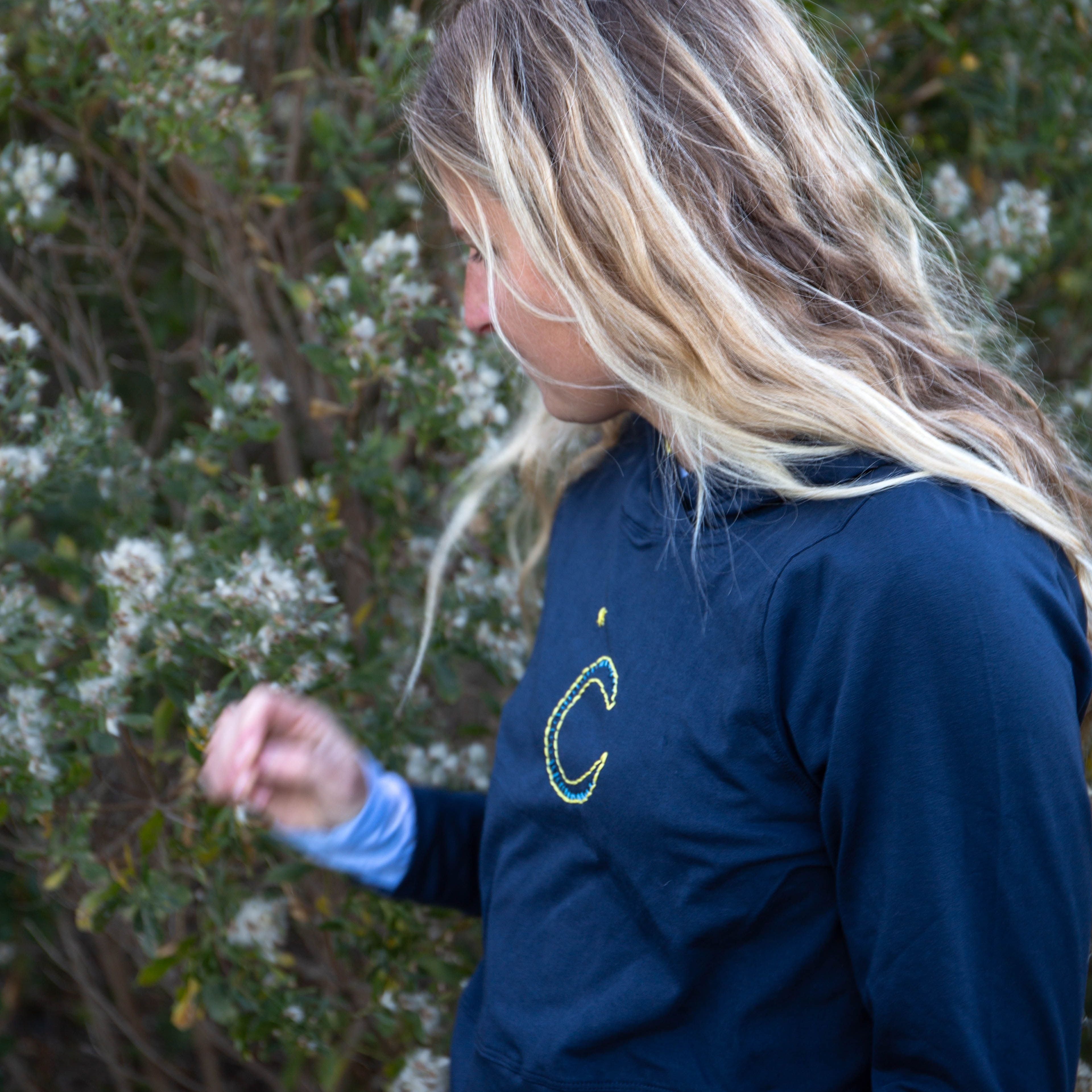 Girl wearing a Yellowberry navy downtown hoodie with yellow embroidery. She is picking a flower.