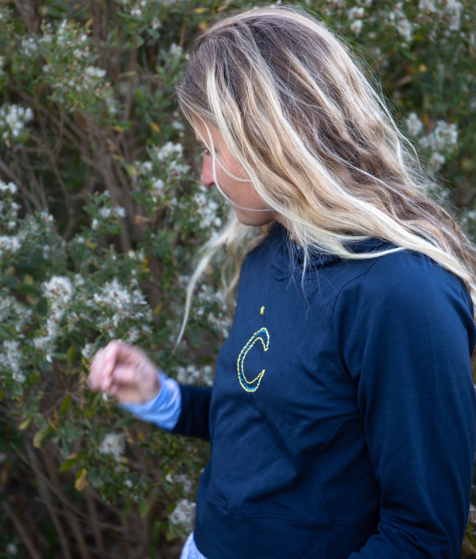Girl wearing a Yellowberry navy downtown hoodie with yellow embroidery. She is picking a flower.