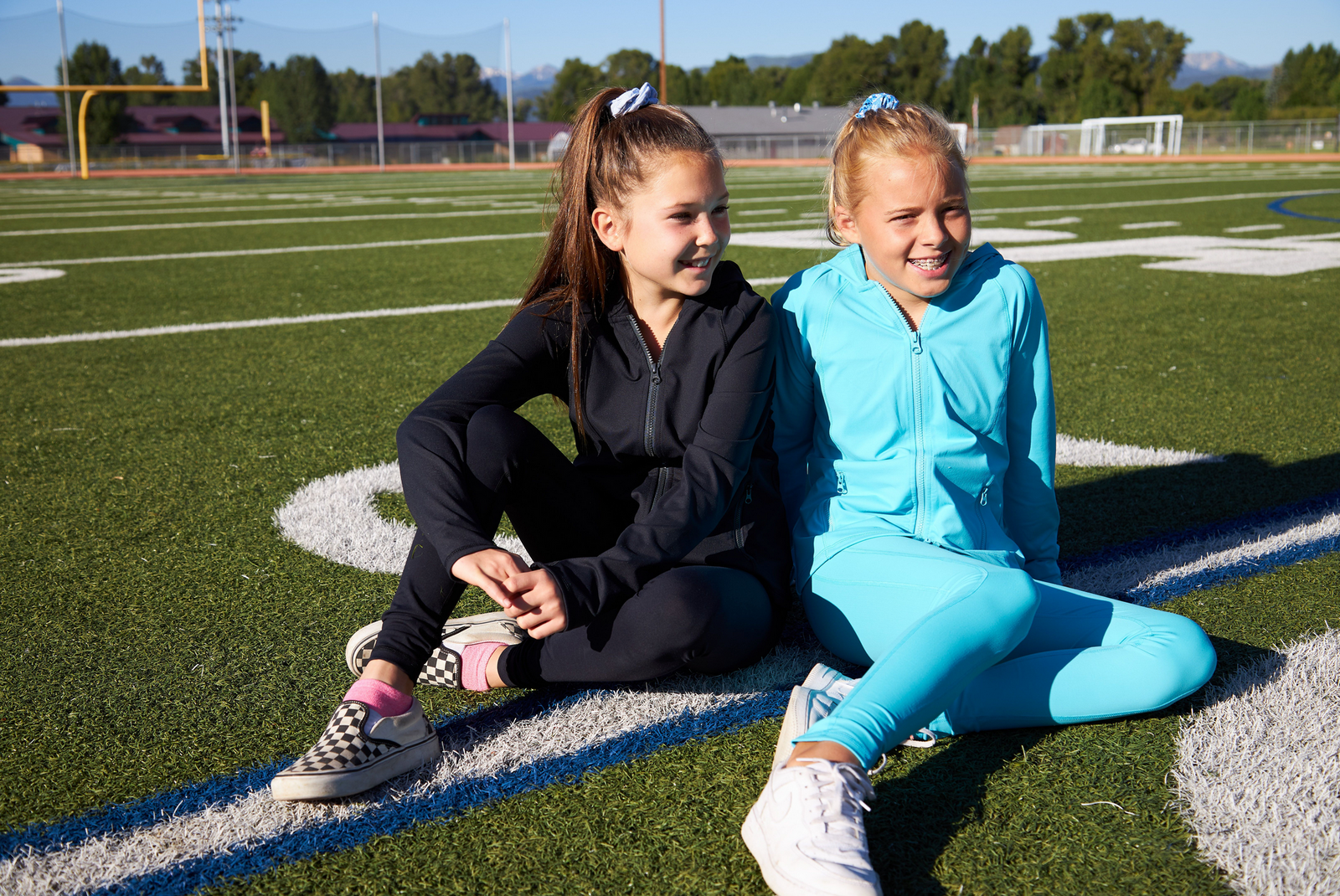 Two girls sitting on an athletic field. One is wearing matching black hoodie and black leggings. The other girl is wearing a matching blue hoodie with blue leggings. Both are athletic matching sets.