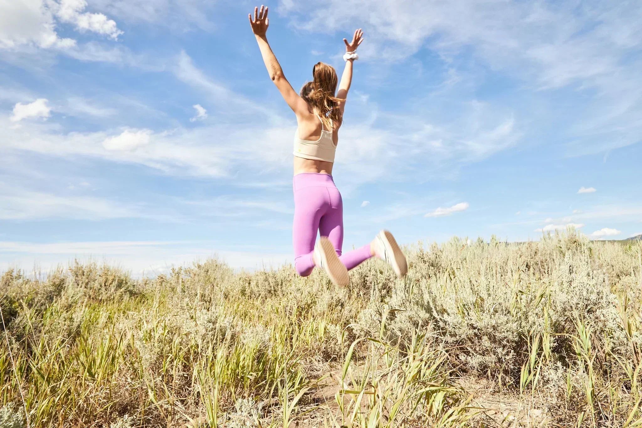 Girl jumping in a meadow with a blue sky behind her. She is wearing a Yellowberry bra and Yellowberry leggings.