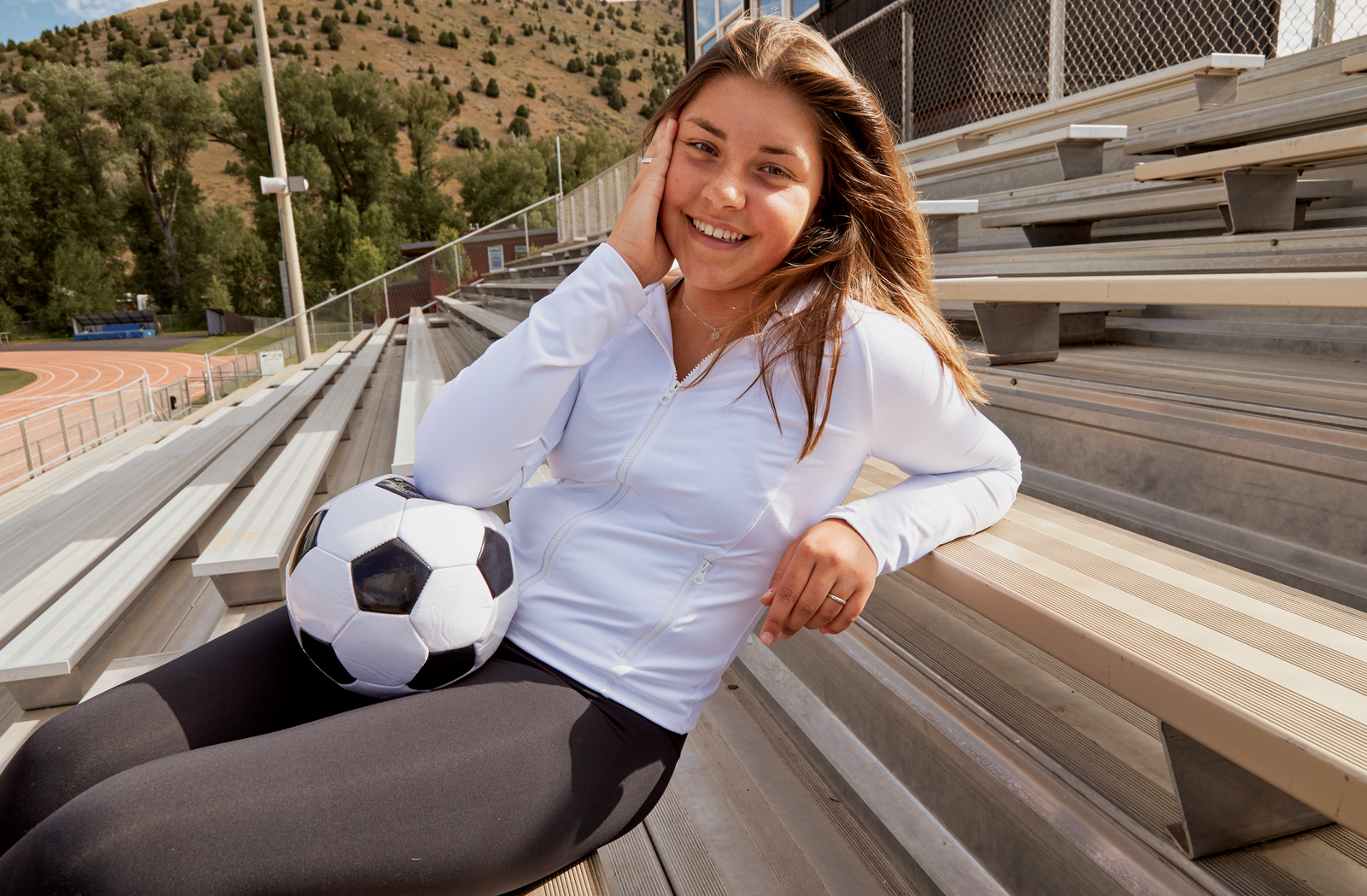 Girl leaning against metal bleachers, holding a soccer ball and smiling in the sun at the camera