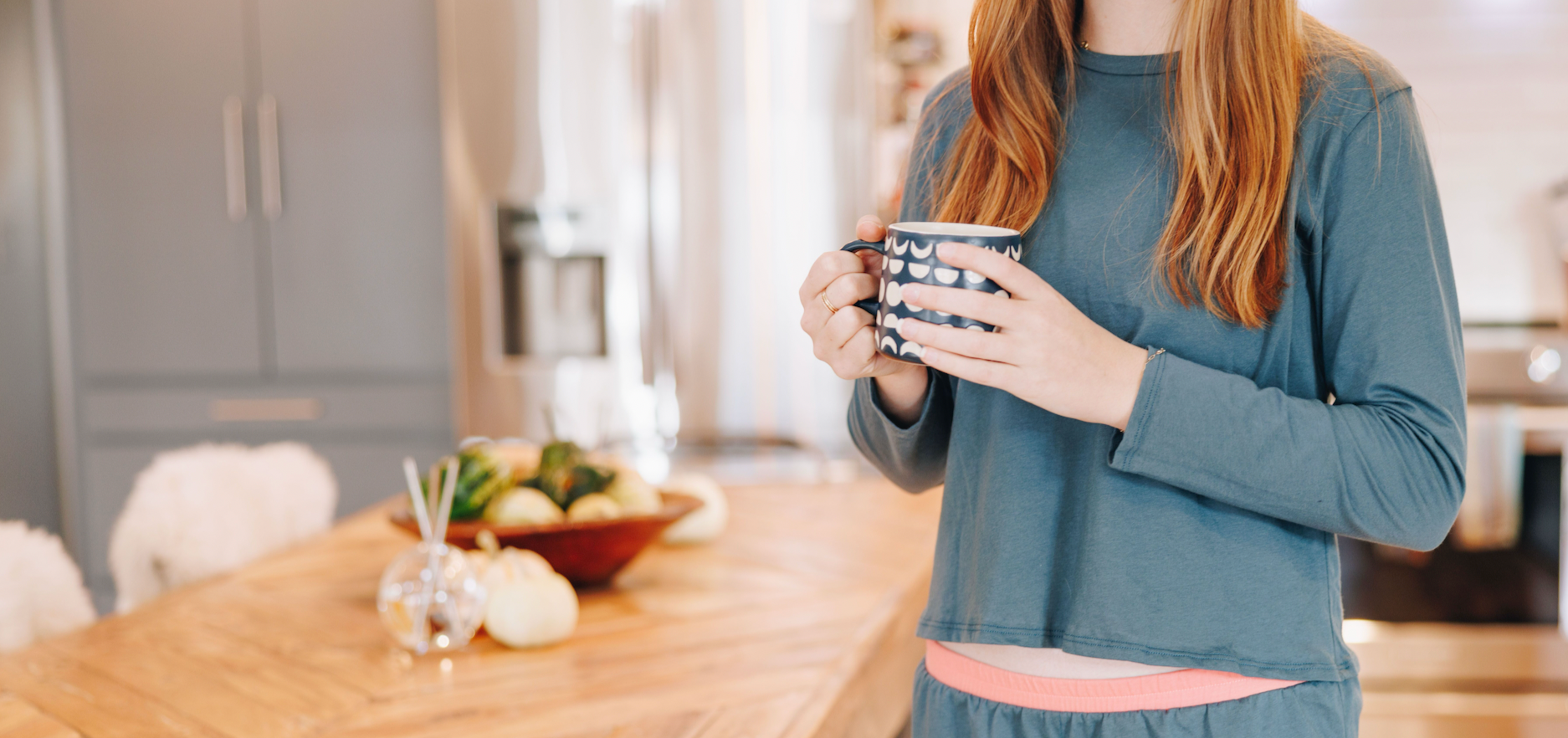 Girl wearing a blueish grey long sleeve t-shirt while holding a cup of tea in a kitchen