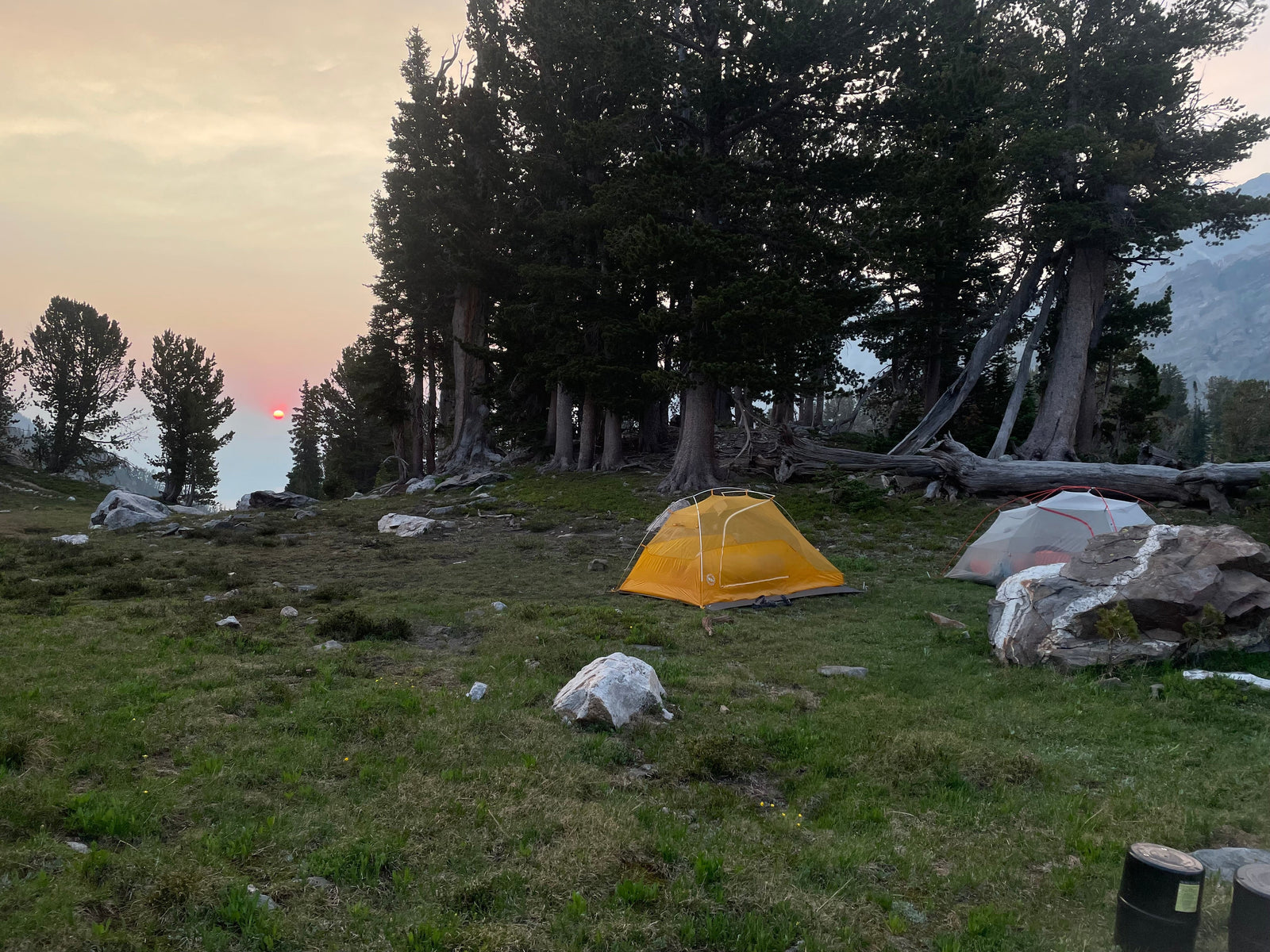 A yellow and white tent are set up on a mountainside. It is the early morning with the sun coming up in the distance.