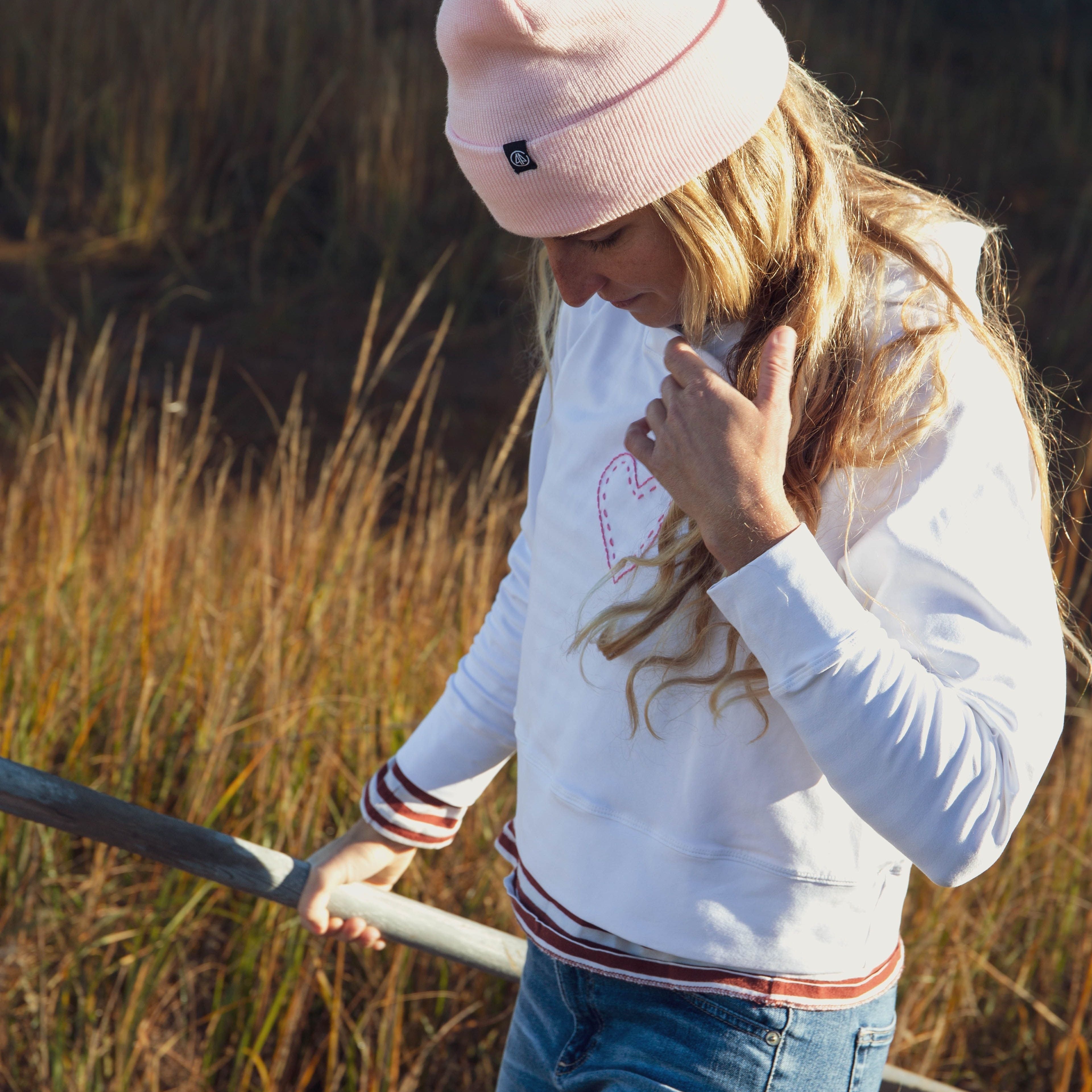 Girl standing in grass, pulling her hair behind her shoulders. She is wearing a Yellowberry Downtown Hoodie and styled it with everyday clothes and a pink hat.