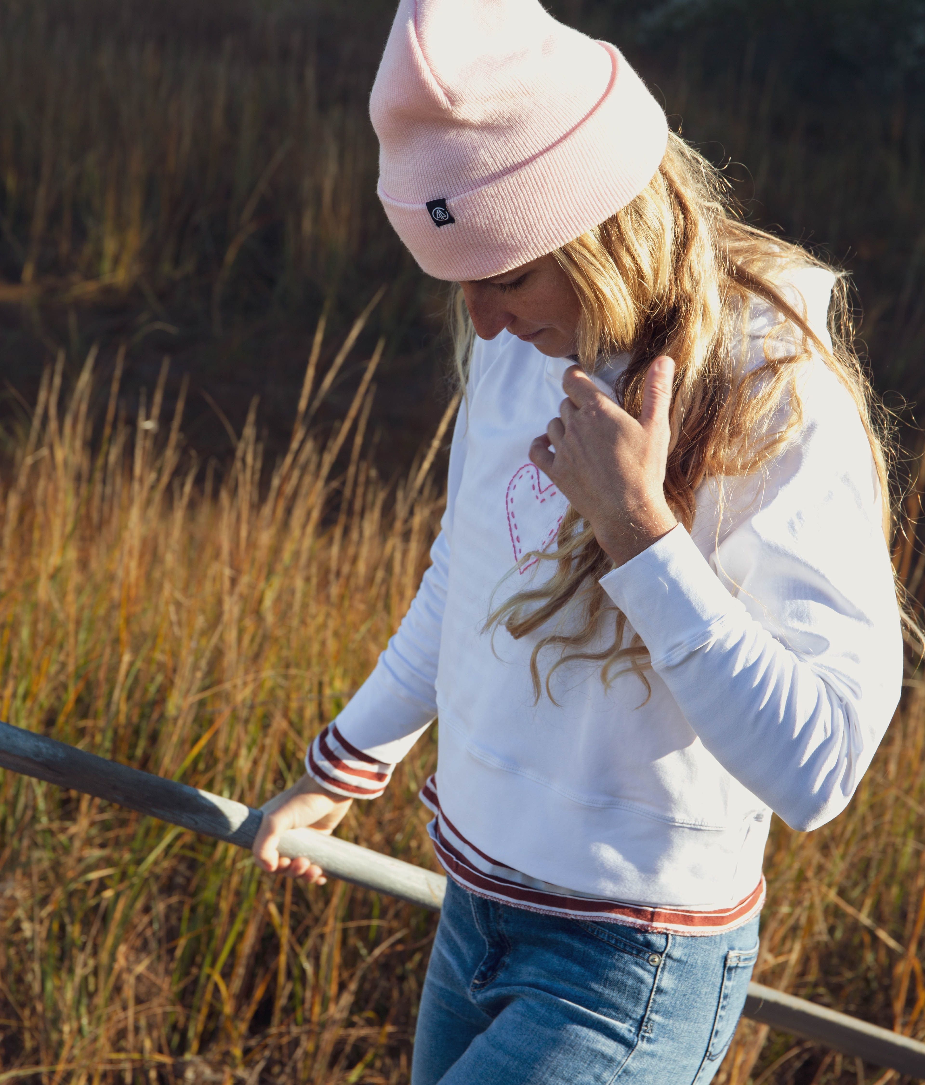 Girl standing in grass, pulling her hair behind her shoulders. She is wearing a Yellowberry Downtown Hoodie and styled it with everyday clothes and a pink hat.