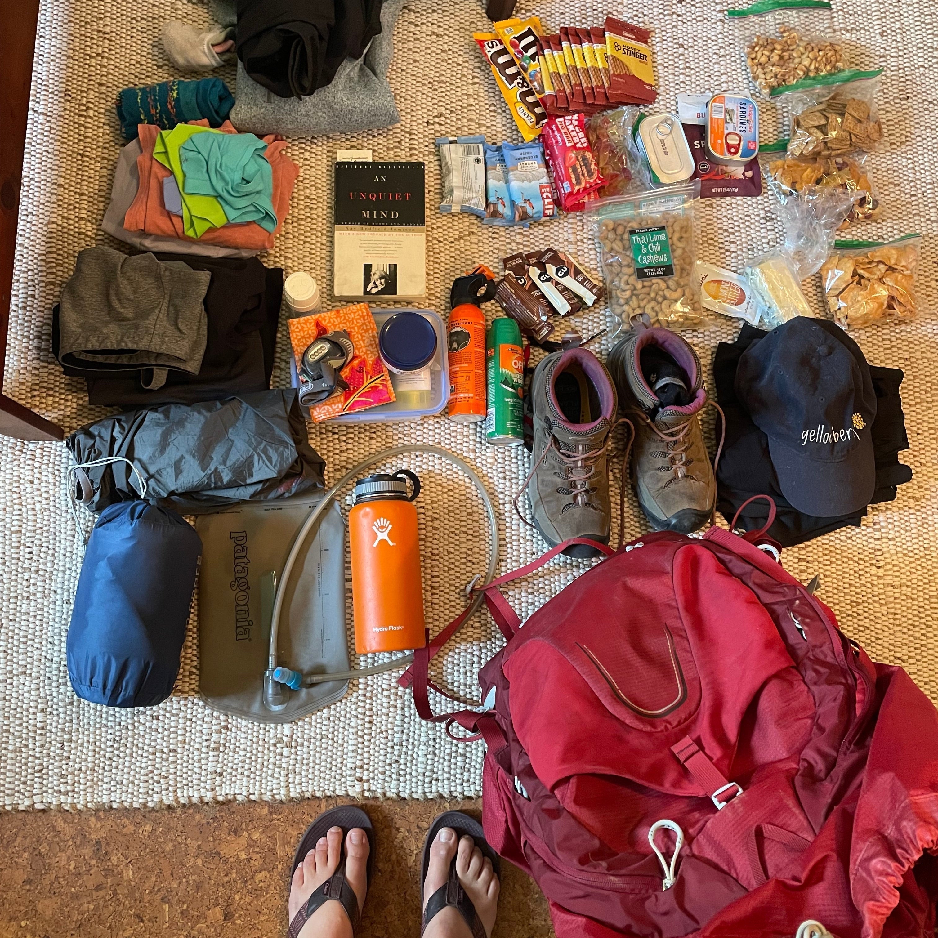 An assortment of camp gear laid out on the floor. There are shoes, hiking boots, bug spray, water filters, day packs, books, hates, clothes, sleeping bags, a backpacking packpack and more.