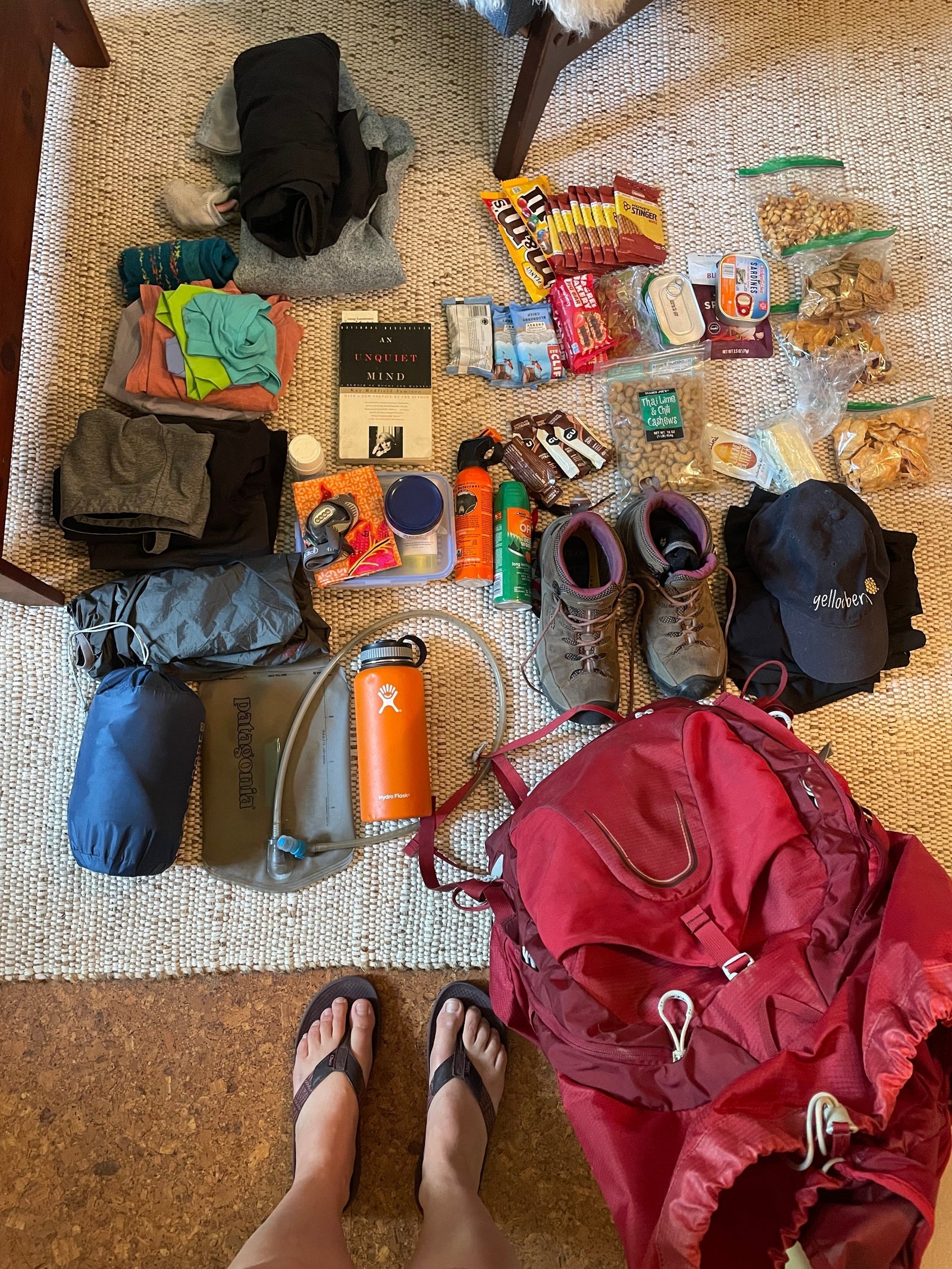 An assortment of camp gear laid out on the floor. There are shoes, hiking boots, bug spray, water filters, day packs, books, hates, clothes, sleeping bags, a backpacking packpack and more.