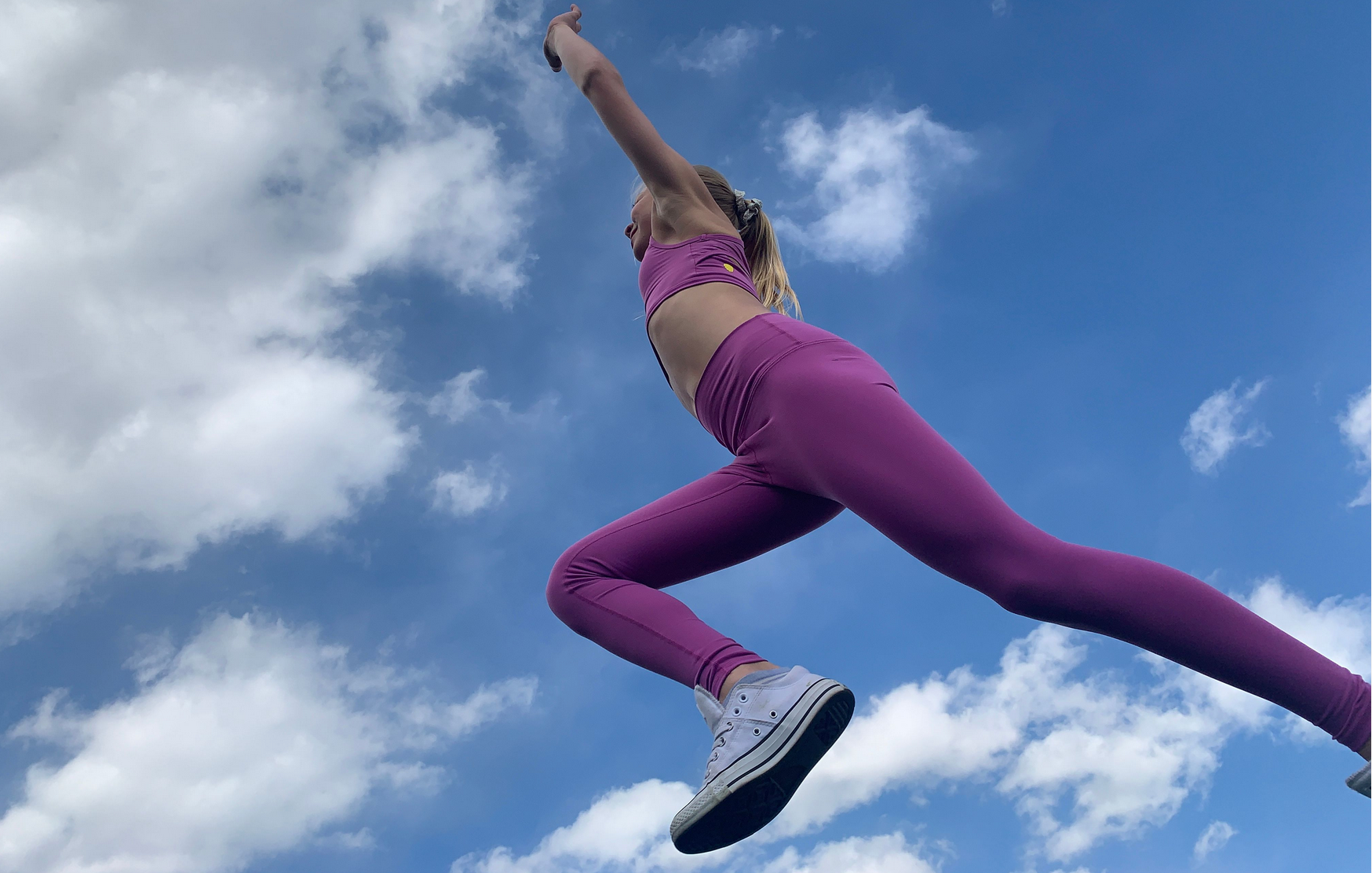 Girl wearing a purple crop top and leggings set. She is jumping up, over the camera with the blue sky behind her
