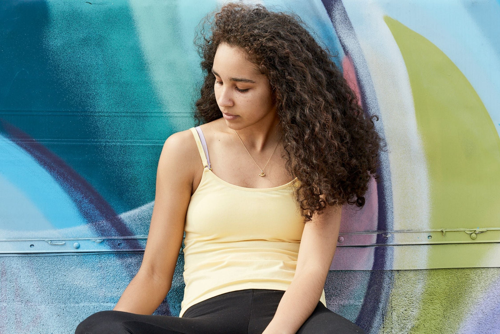 Girl sitting down, leaning against a colorfully painted wall. She is wearing a Yellowberry yellow cotton camisole and leggings.