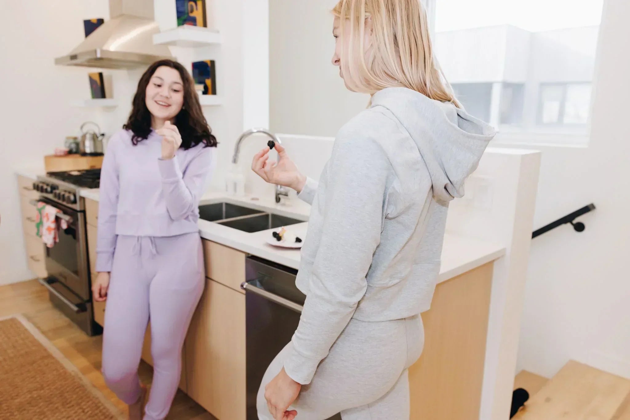 Two girls standing in the kitchen at a counter in Yellowberry lounge sets. They are eating berries, and one is wearing a lilac colored lounge set and the other is in a grey colored lounge set.