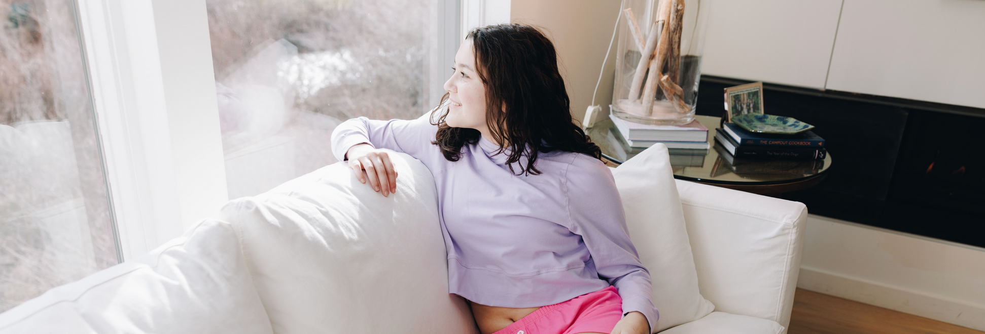 Girl wearing a light purple cotton hoodie and bright pink shorts while sitting on a white sofa next to a large window