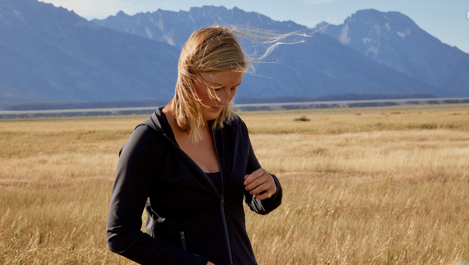 Girl standing in front of a mountain range in the distance in a golden meadow. She is looking down and zipping up her black Yellowberry hoodie.
