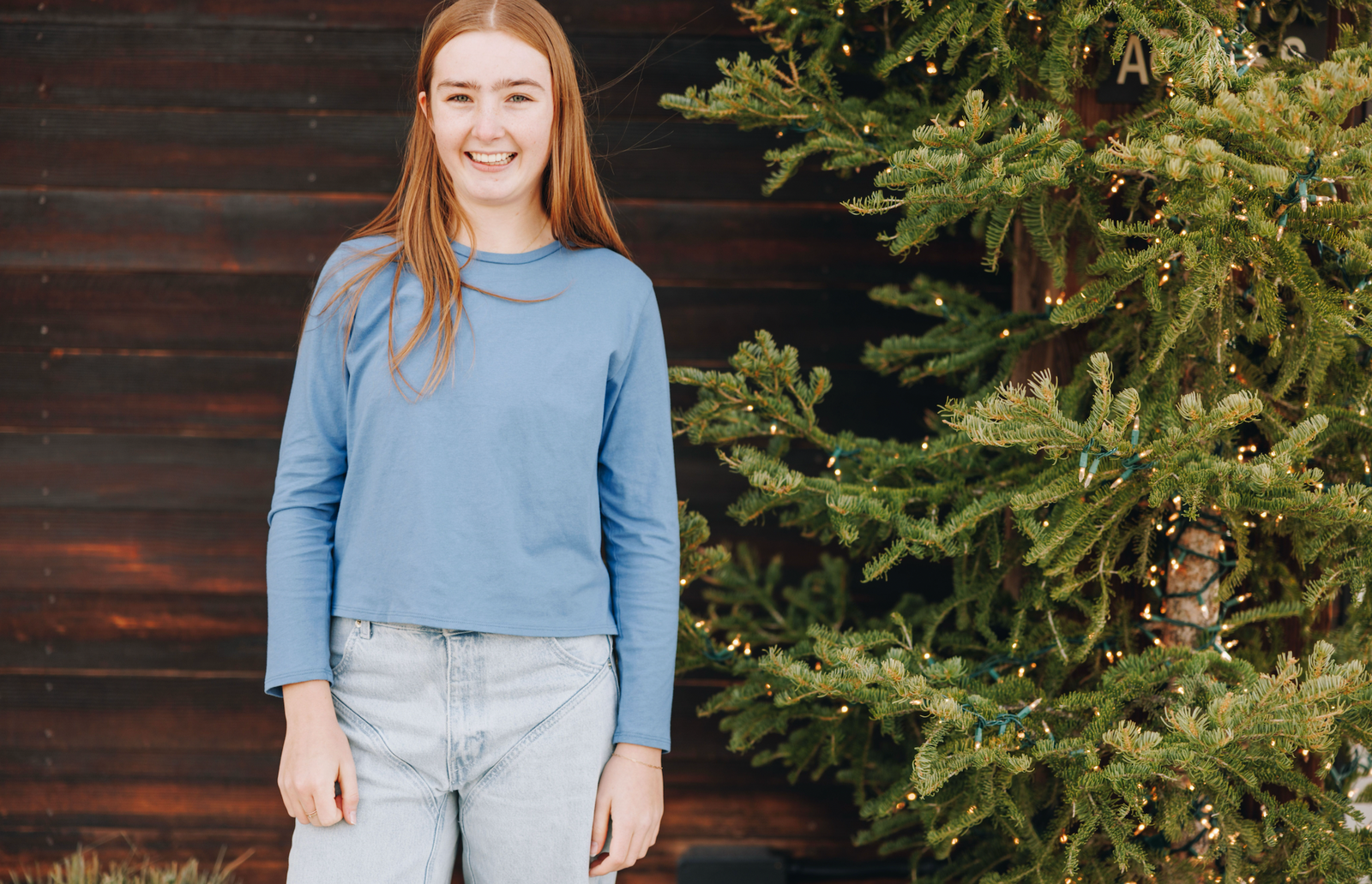 Girl smiling wearing a blue long sleeve t-shirt against a tree with lights and wooden wall