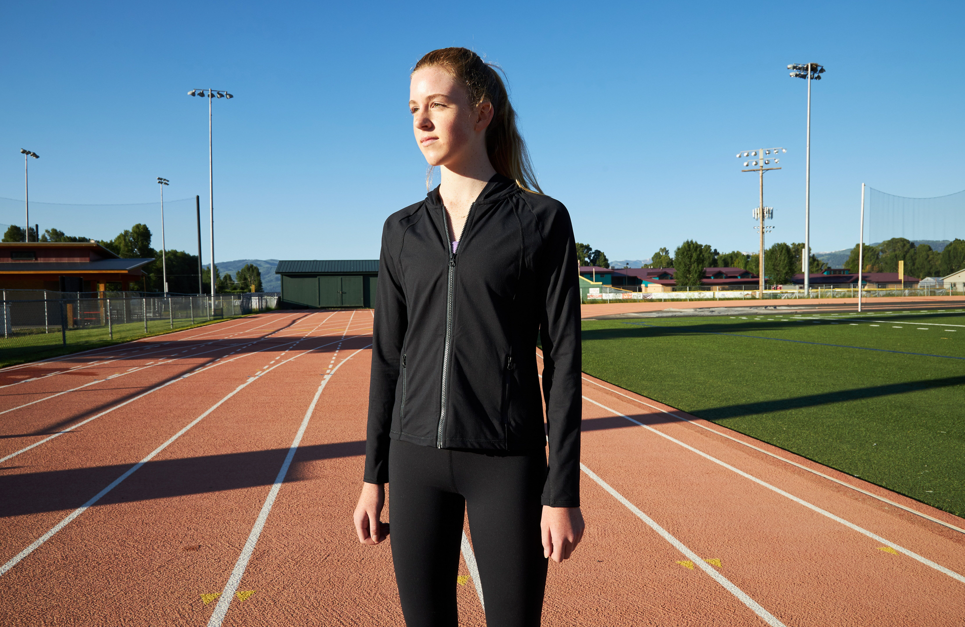 Girl standing on an organge running track. Her hair is in a ponytail and she is wearing a black hoodie with black leggings.