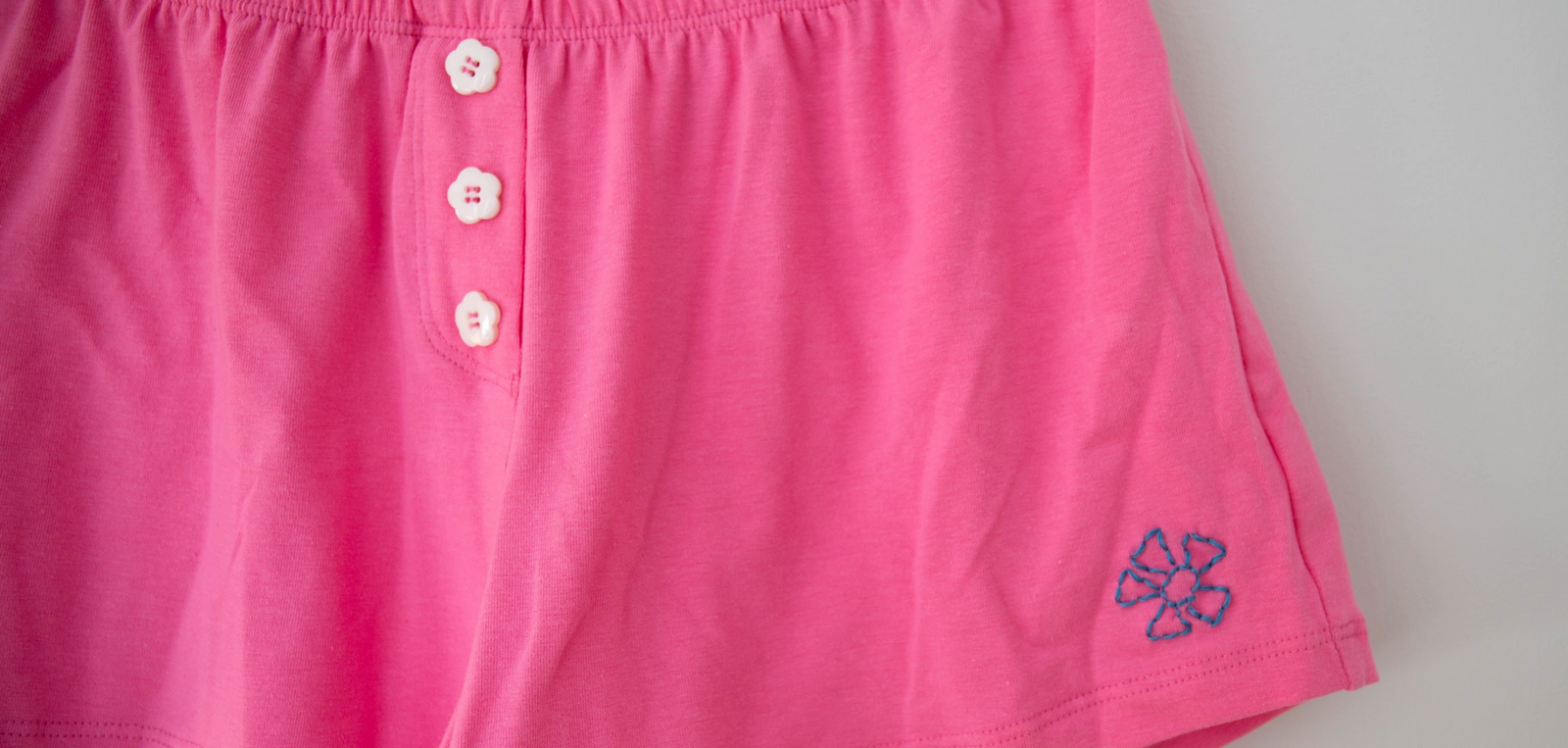 Pink cotton shorts with a blue embroidered flower and three white buttons