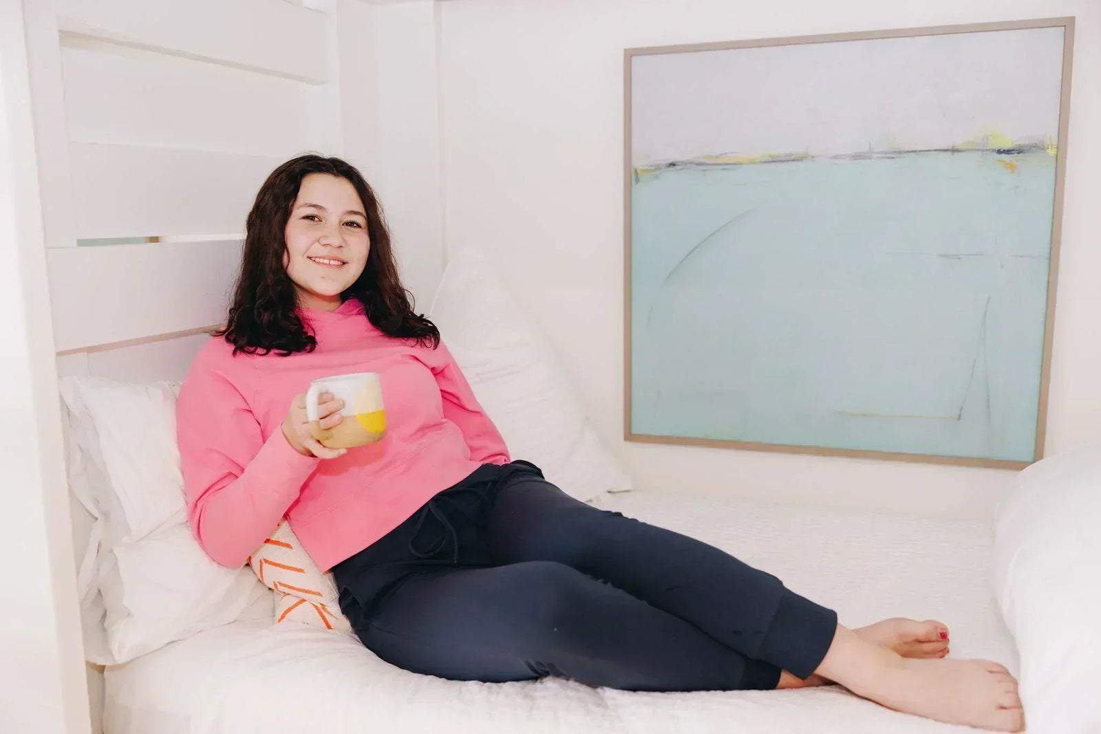 Girl lounging on a bed wearing a hot pink Yellowberry Downtown Hoodie and navy Uptown Jogger lounge pants. She is holding a warm drink cup in her hand.