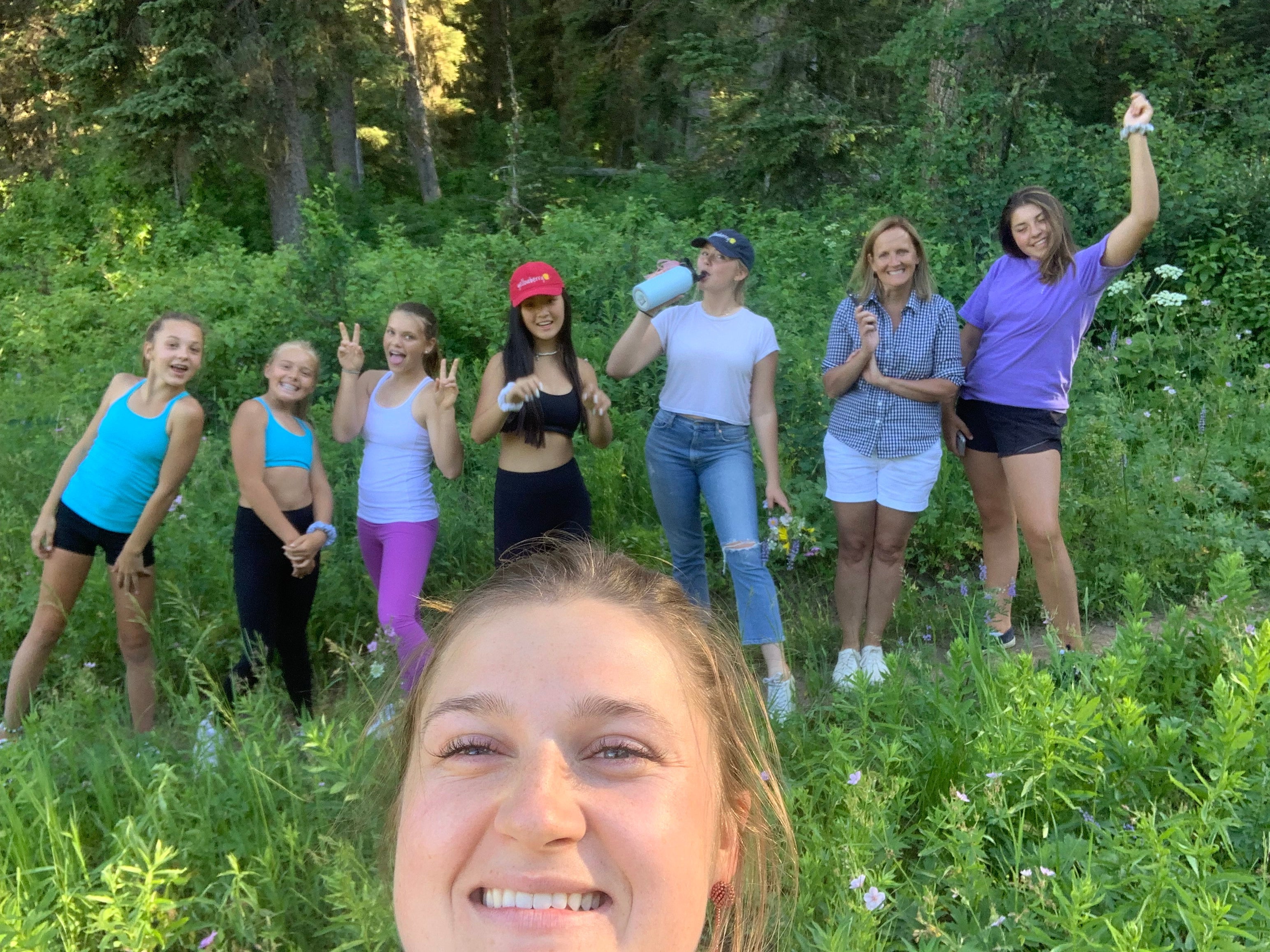 Girl taking a selfie smiling with a line of girls on the trail outside behind her - everyone is smiling, on a summer day