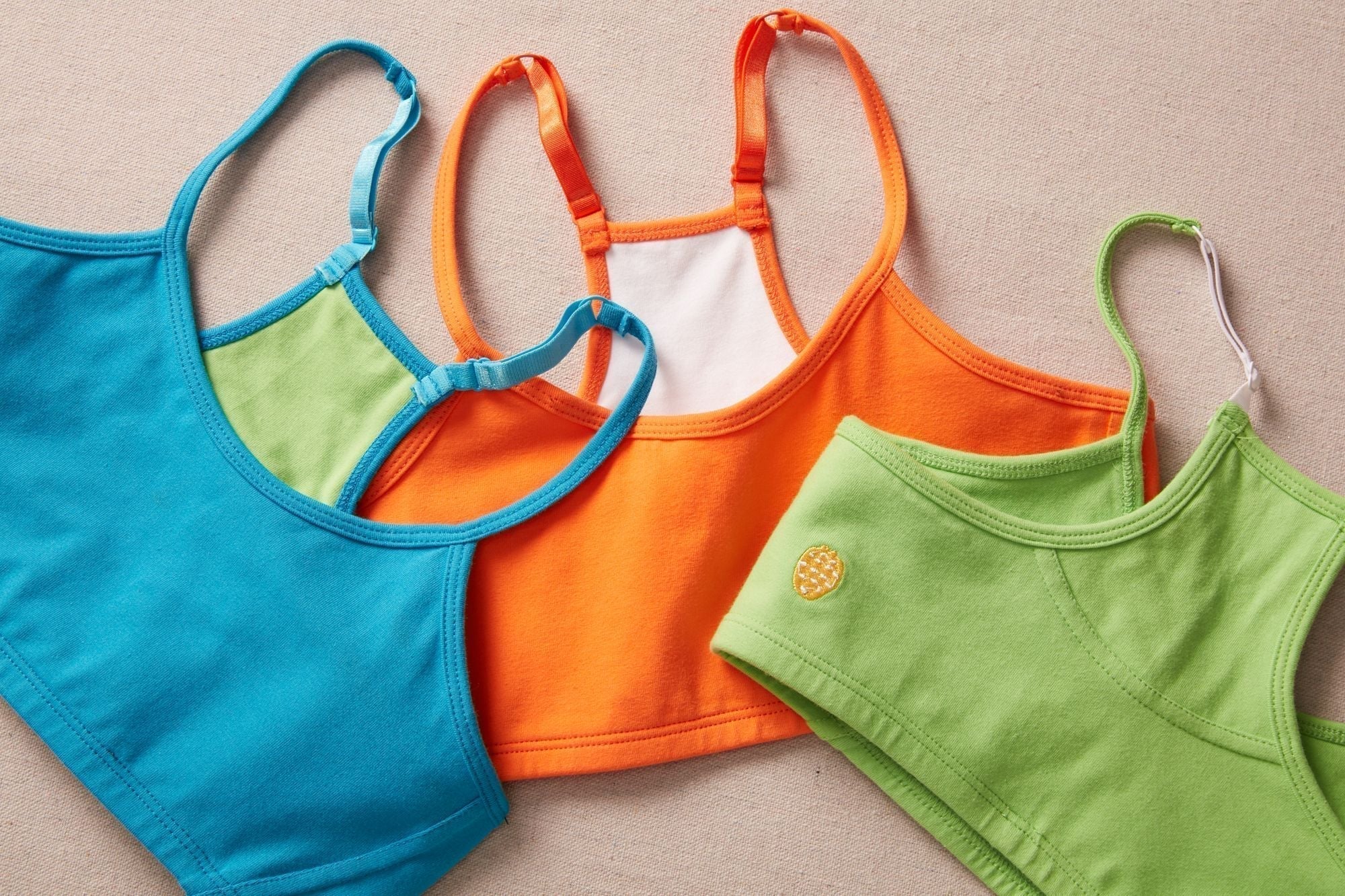 Three colorful cotton bras on a light linen background. There is a blue bra, an orange bra, and a green bra.