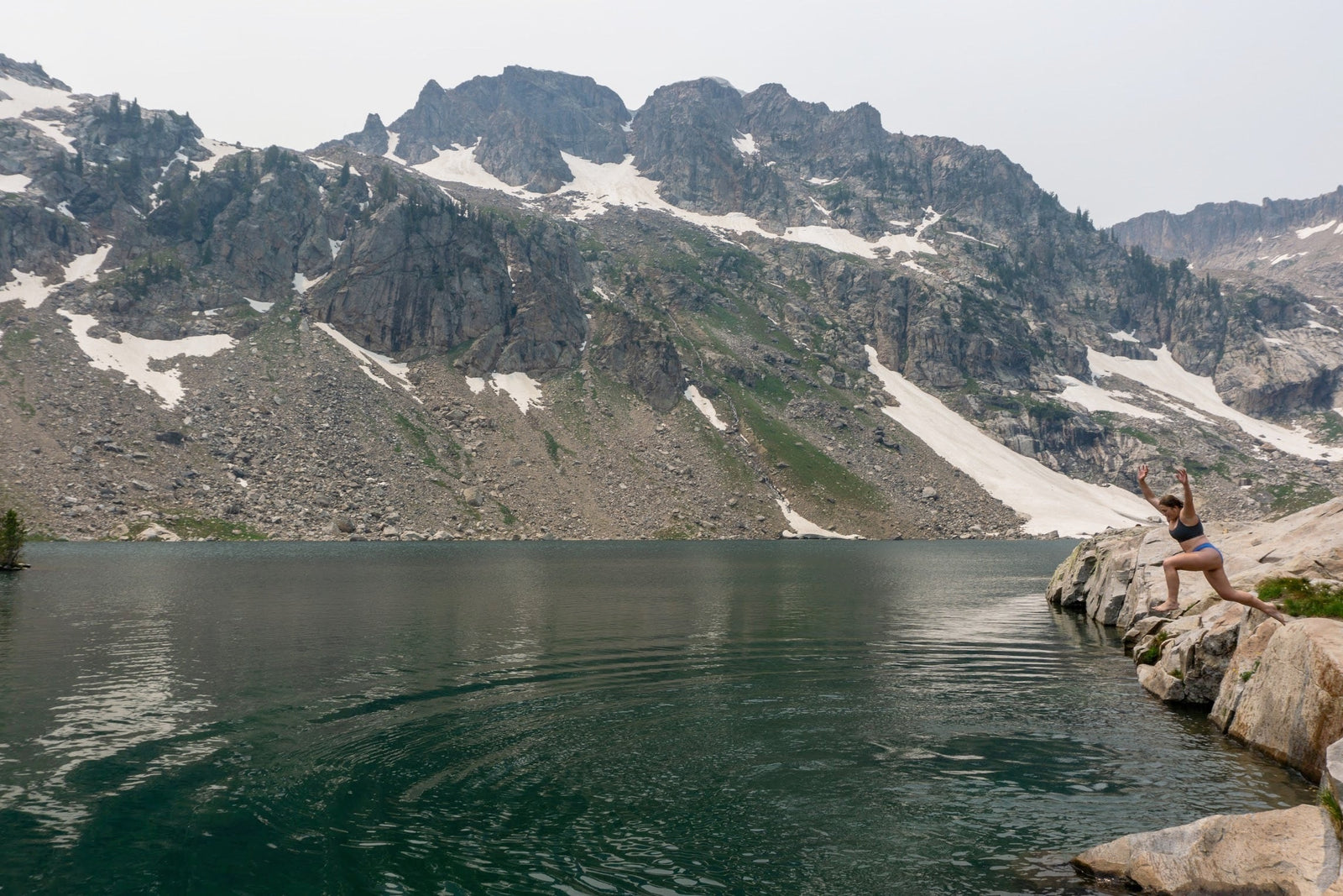 Girl jumping off of a rocky edge into a mountain lake. The lake is surrounded by glacier snow and rocky mountains. She is smiling and the water must be cold! 
