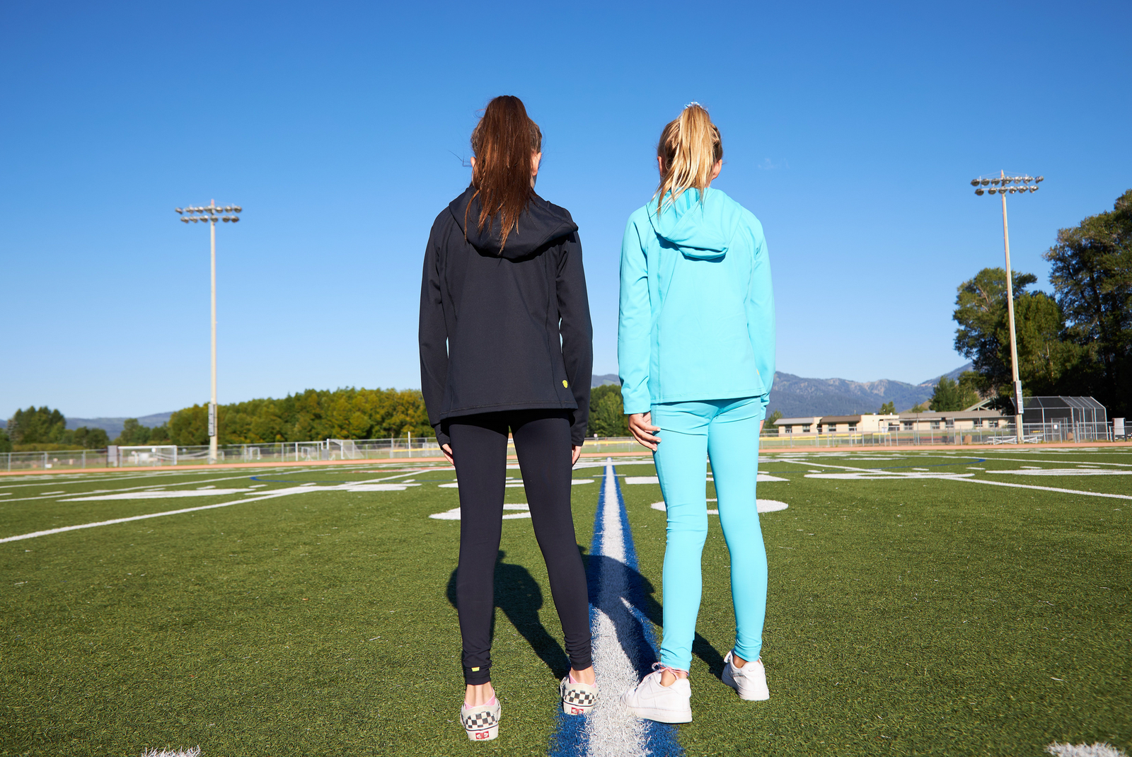 Two girls standing on an athletic field. They are each wearing matching sets - a hoodie with matching leggings. One is wearing all black, he other is wearing all light blue.