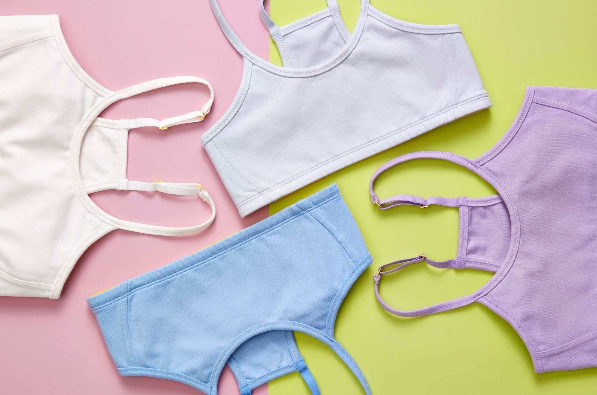 Four Yellowberry Bras styled on a two-color background. The cotton bras are white, light blue, pale blue, and lighter purple. The background is green and pink.